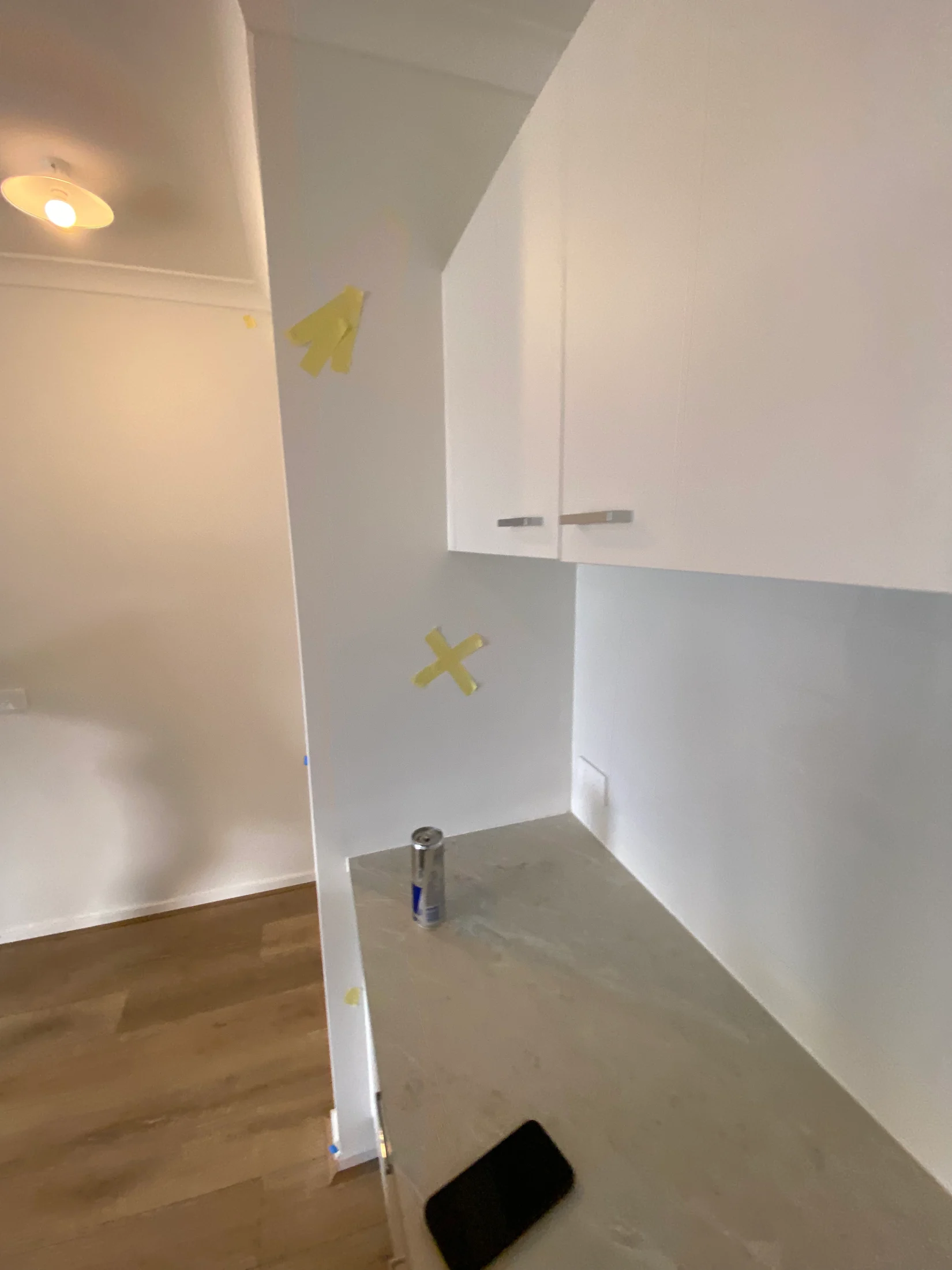 White kitchen with yellow tape on walls, a countertop with a phone and drink can. Wooden floor, light fixture in background.