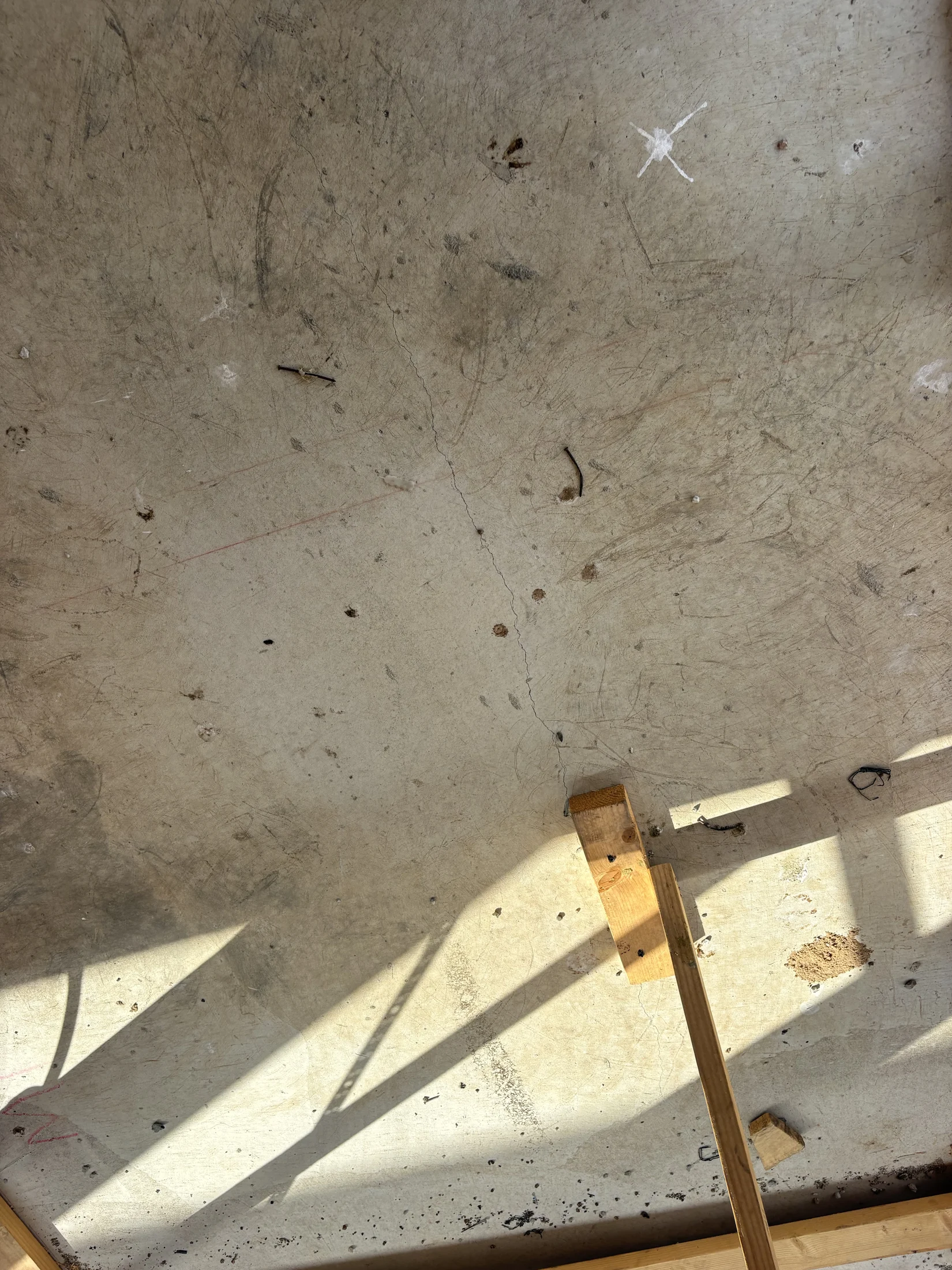 Sunlight casts shadows of wooden beams on a scratched concrete floor. Debris and marks are scattered, creating a rugged, industrial feel.