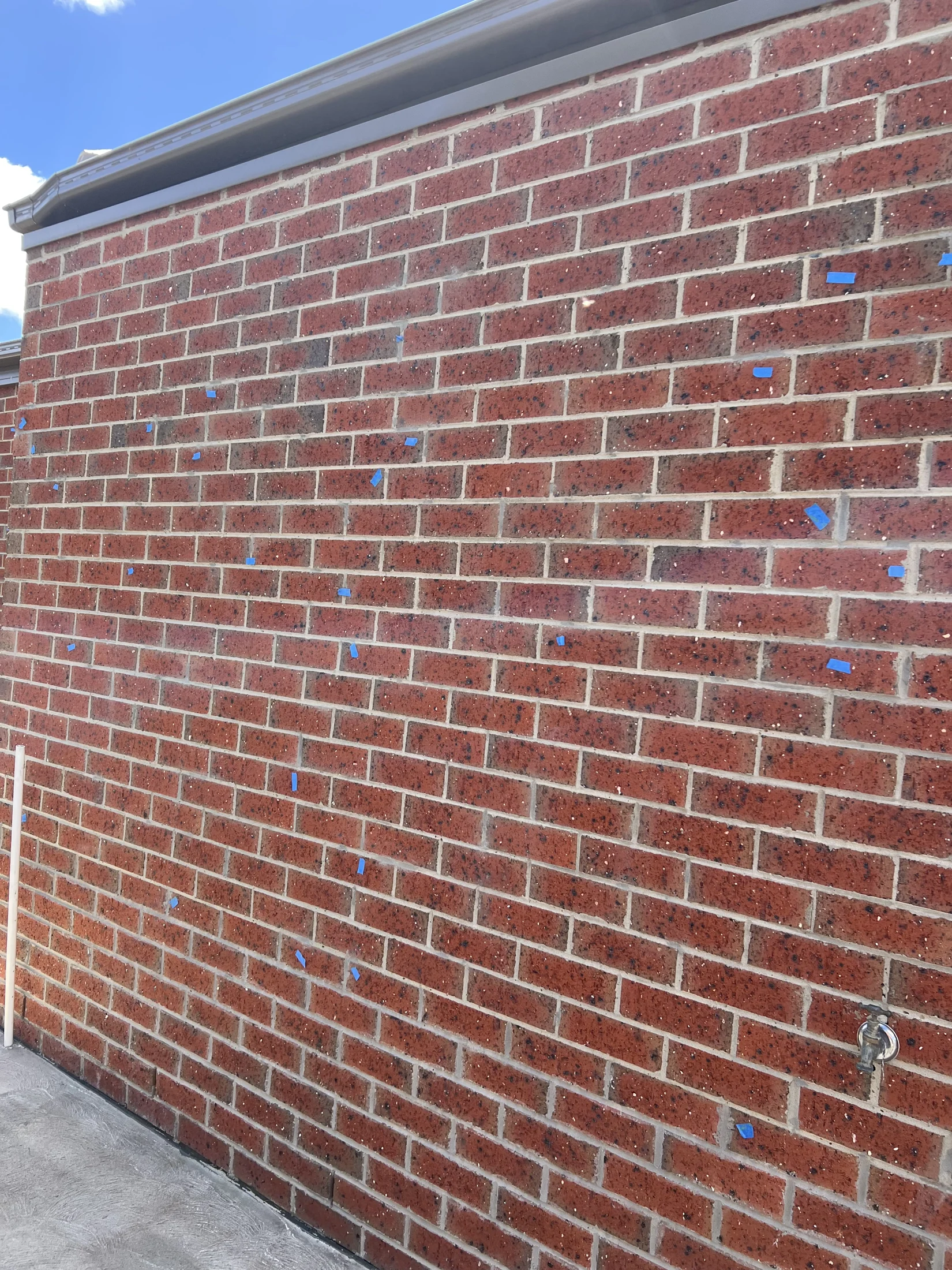 Red brick wall with scattered blue tape pieces, metal fixture on right. Overcast sky above and concrete ground below.