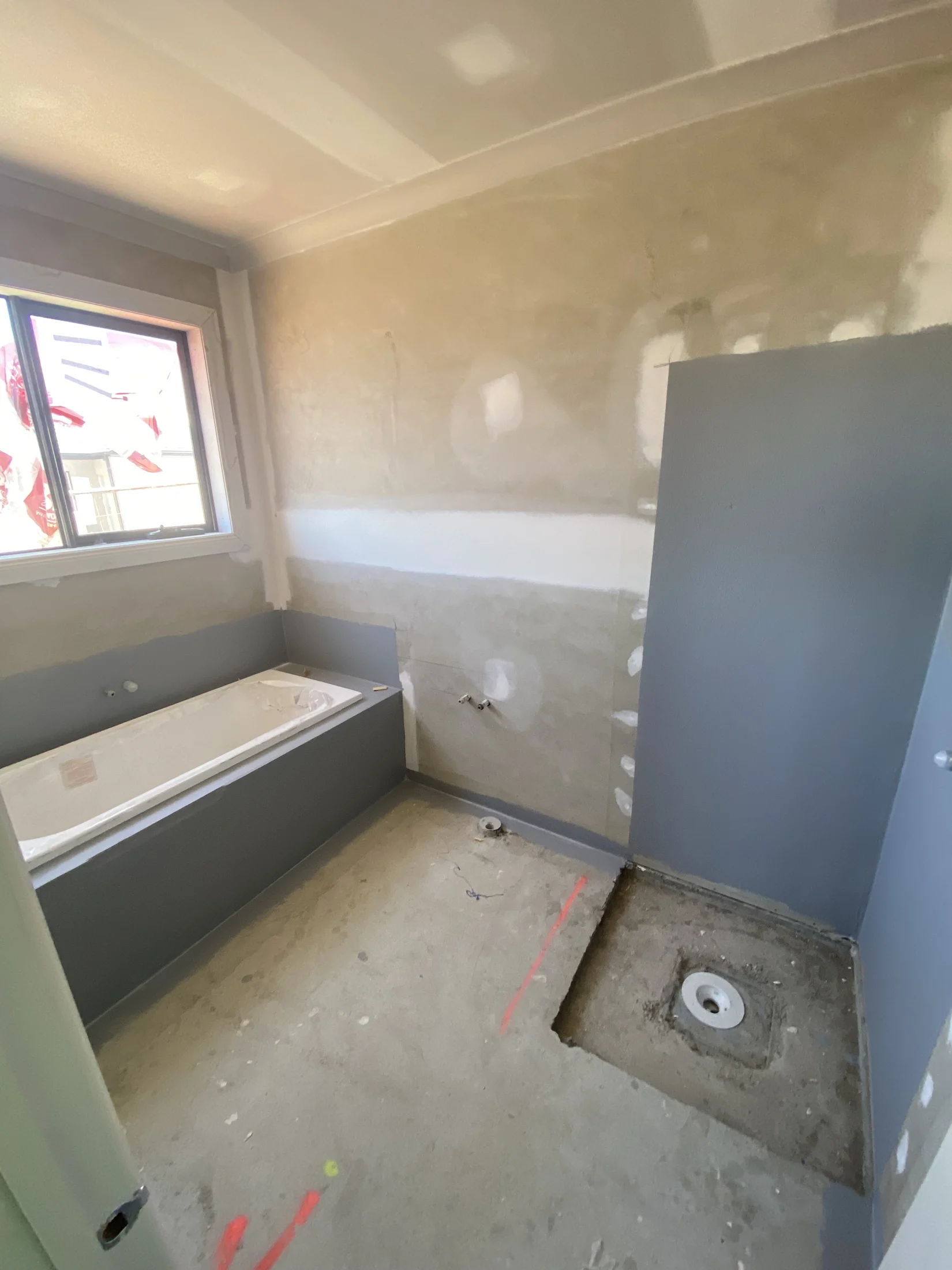 Bathroom under renovation with unfinished walls and a bathtub. Exposed plumbing and concrete floor. Window showing construction outside.