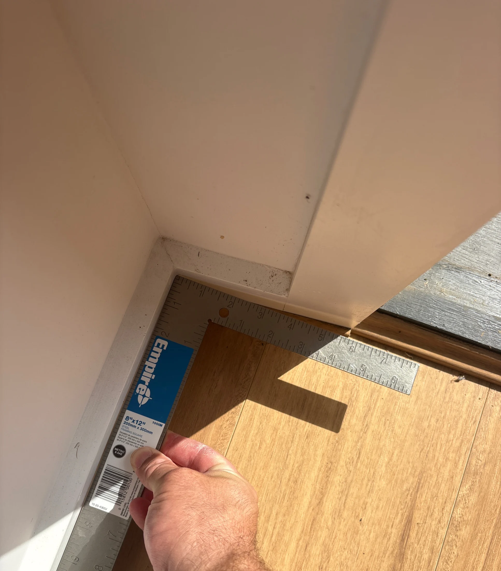 Hand holding an Empire square ruler measures a corner on a light wood floor, adjacent to a white wall. Bright sunlight casts shadows.