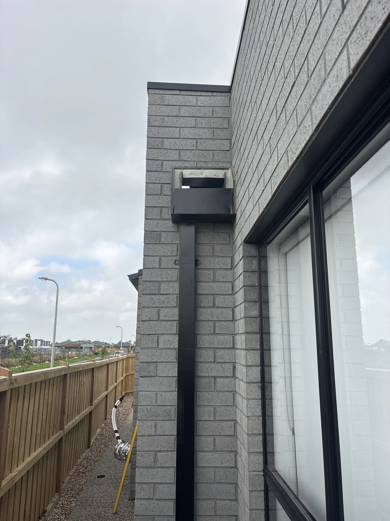 Brick building exterior with gray bricks, black vent pipe, and large window. Wooden fence and cloudy sky in background. Urban setting.