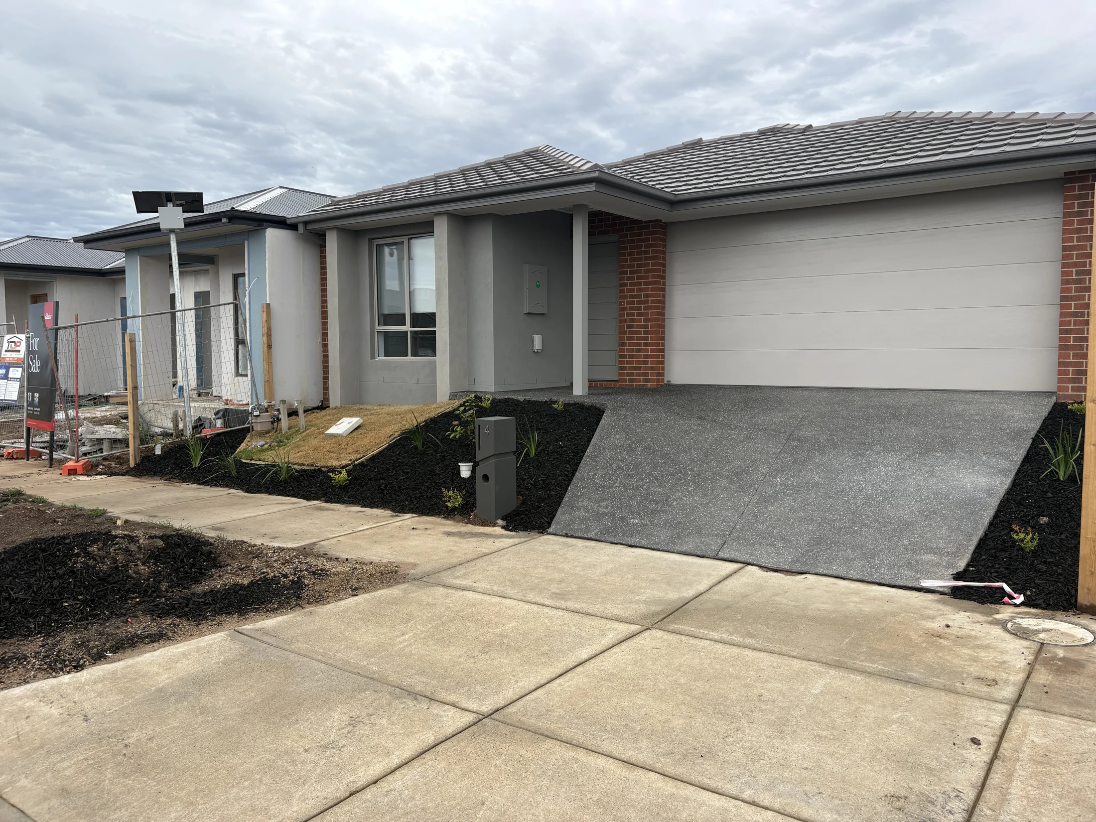 Modern house with gray garage, brick accents, and landscaped yard under cloudy sky. "For Sale" sign visible beside a neighboring house.