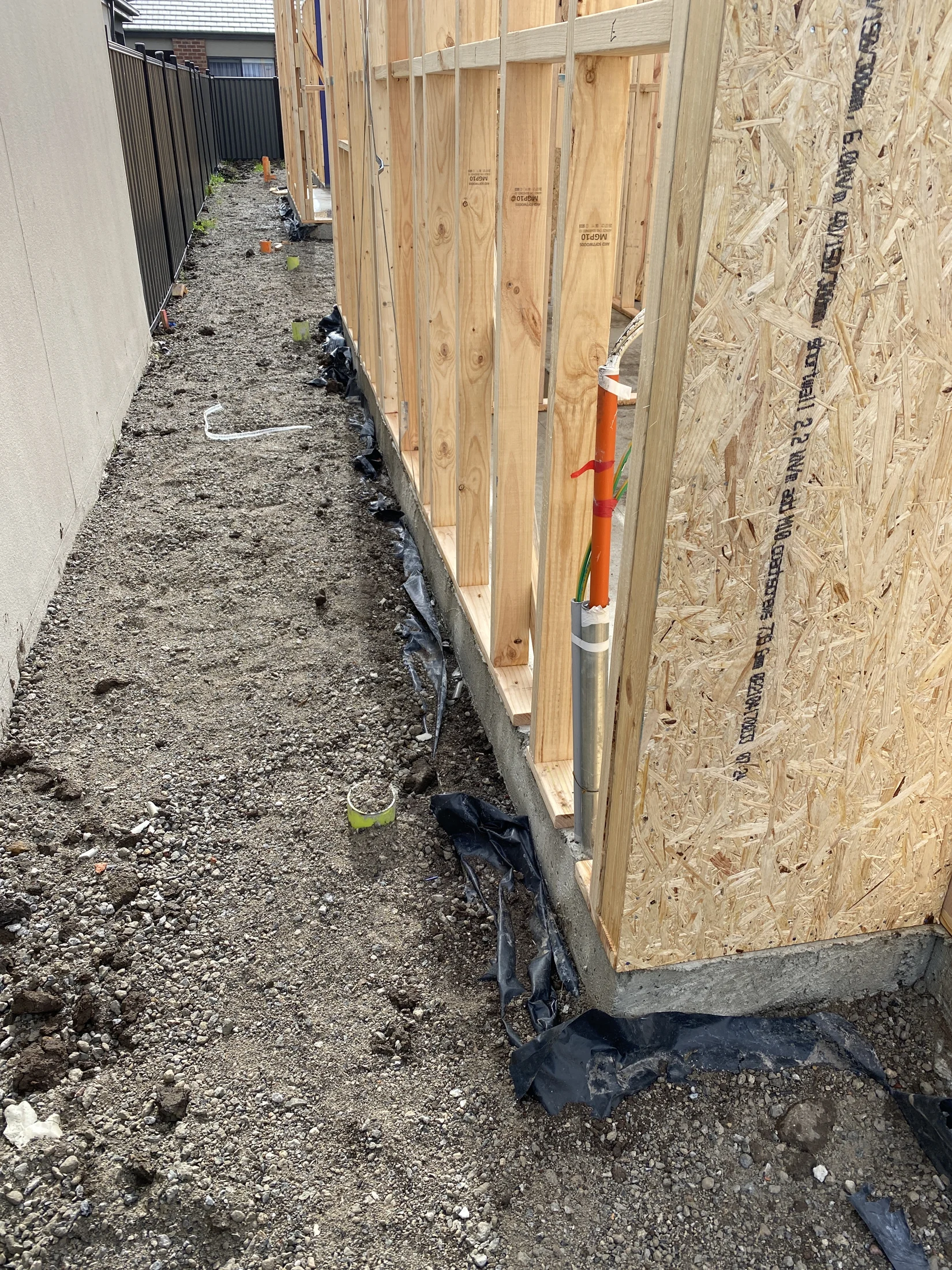 Construction site with wooden wall framing on a gravel path. Exposed pipes and black plastic sheeting visible. Overcast day, muted colors.