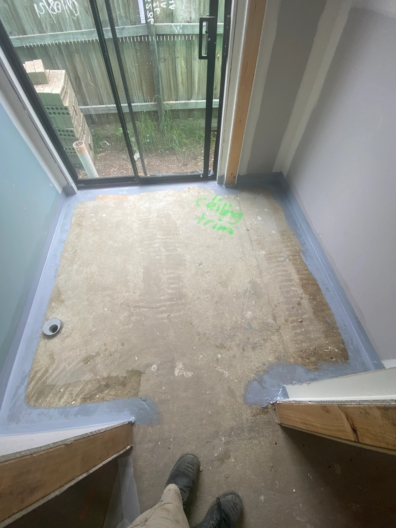 View of a small room with unfinished flooring, blue sealant along edges, and “celling time” in green. Glass door leads to fenced yard.