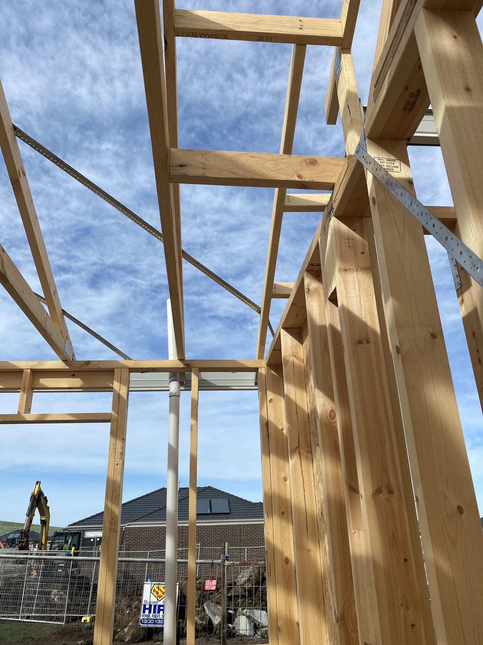 Wooden house frame under blue sky, white clouds; construction site with fence and excavator in background, HIA sign visible.