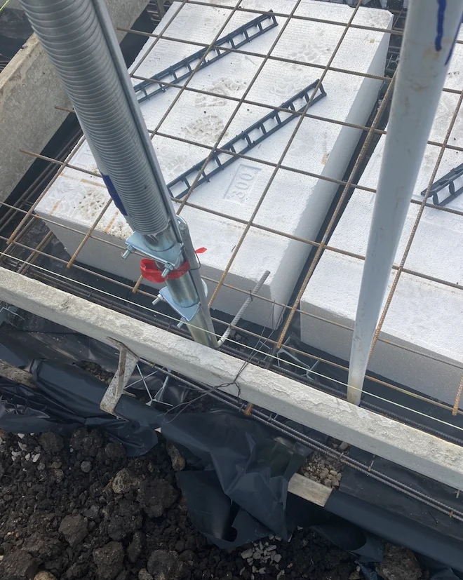 Construction site with white blocks and metal bars on a wire grid. A pole with a red clamp is in the foreground. Soil and black plastic visible.