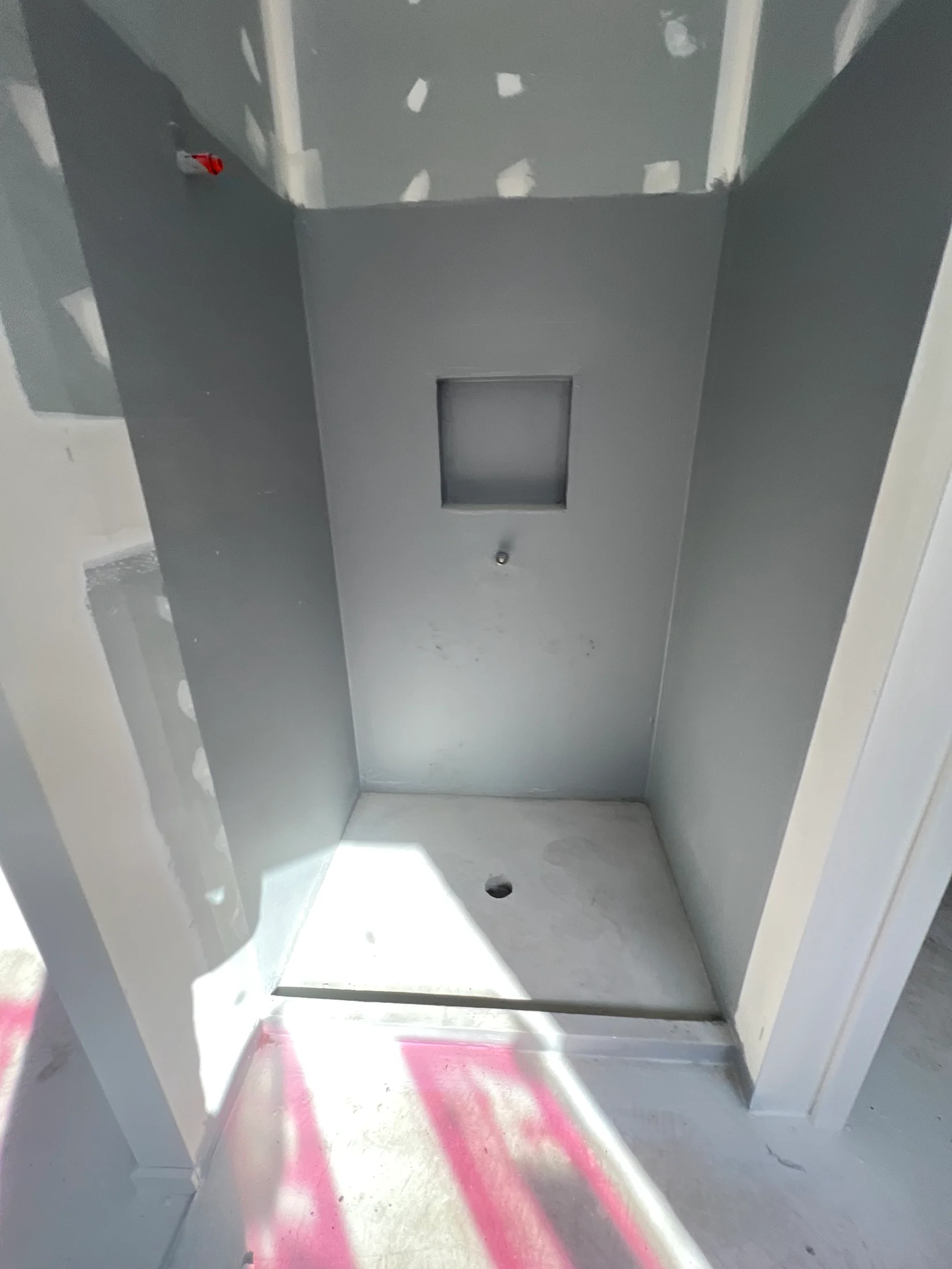A partially constructed shower space with waterproofed walls, a floor drain, and a niche. Sunlight casts shadows, and pink paint marks the floor.