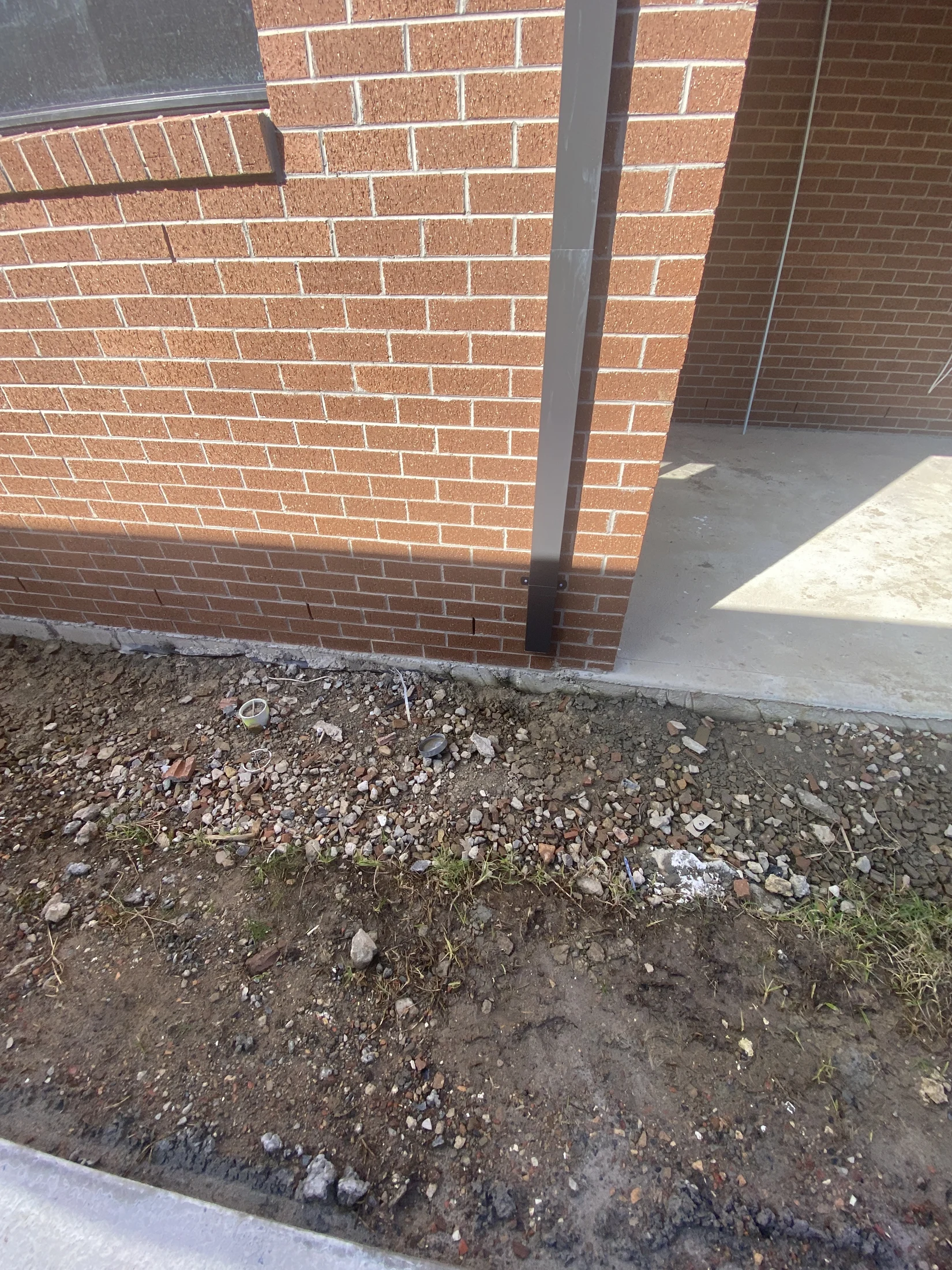 Red brick wall with a vertical metal beam; dirt and scattered rocks in front. Sunlight casts shadows on concrete floor.