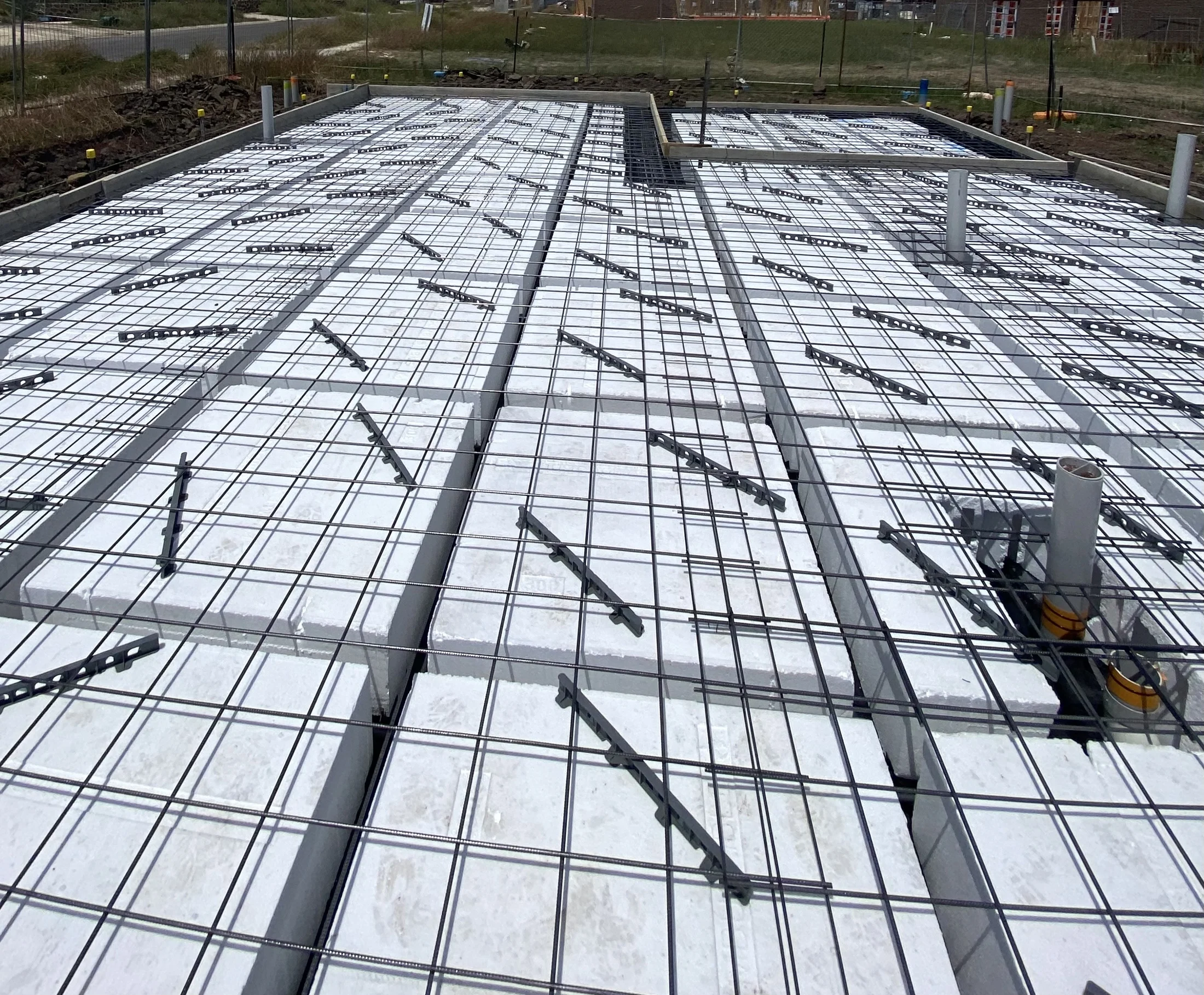 Concrete slab with grid-like steel reinforcement on a construction site, set against a grassy background. Overcast sky.