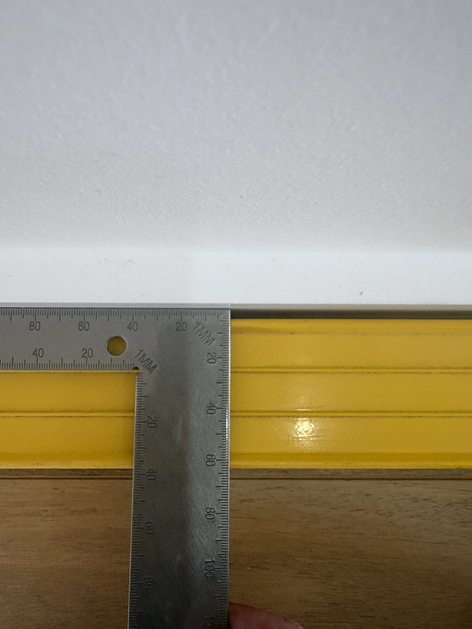 Metal ruler measuring a white wall and yellow trim. The background is wooden, and ruler markings are visible. Calm, precise setting.