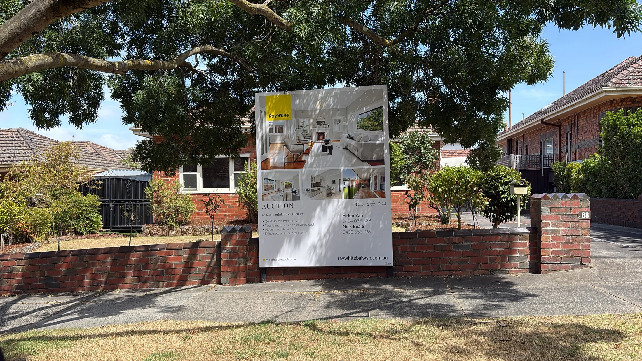 Real estate auction sign outside a brick house with a garden, under a large tree. The sign shows interior photos and contact info.