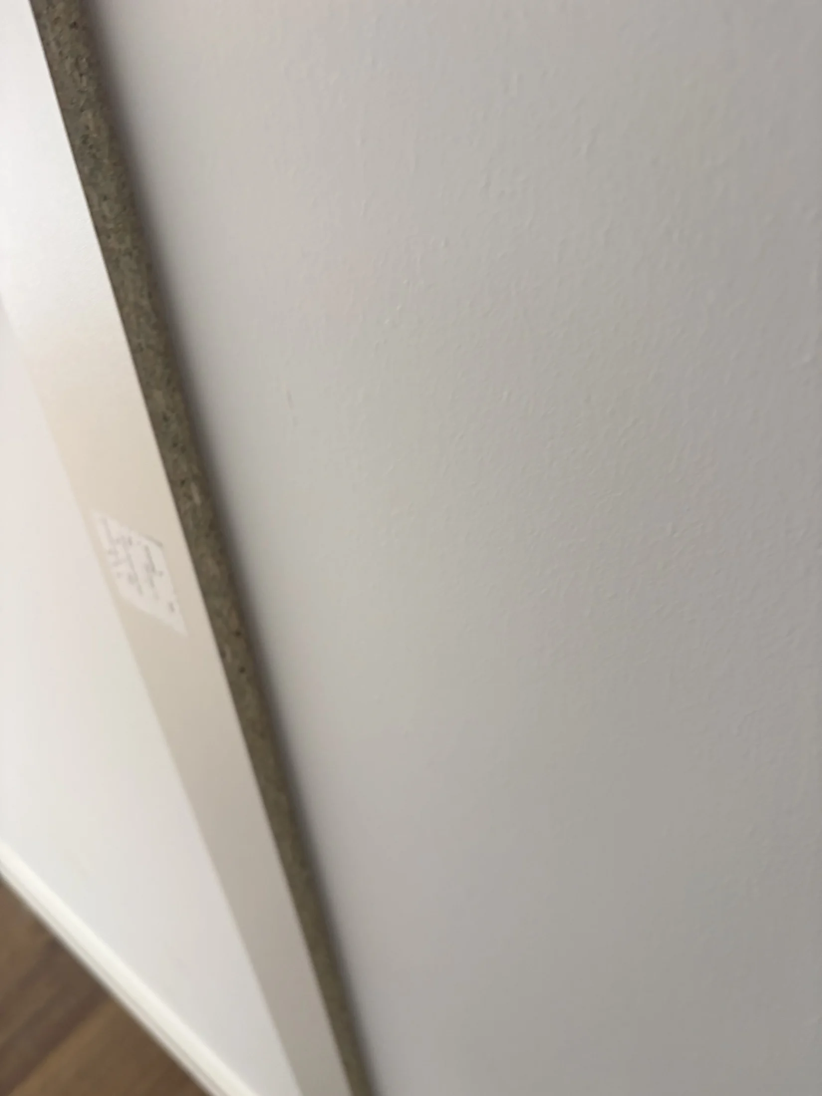 Close-up of a white wall and edge of a painting or picture frame, with a small label visible. Wooden floor in the background.