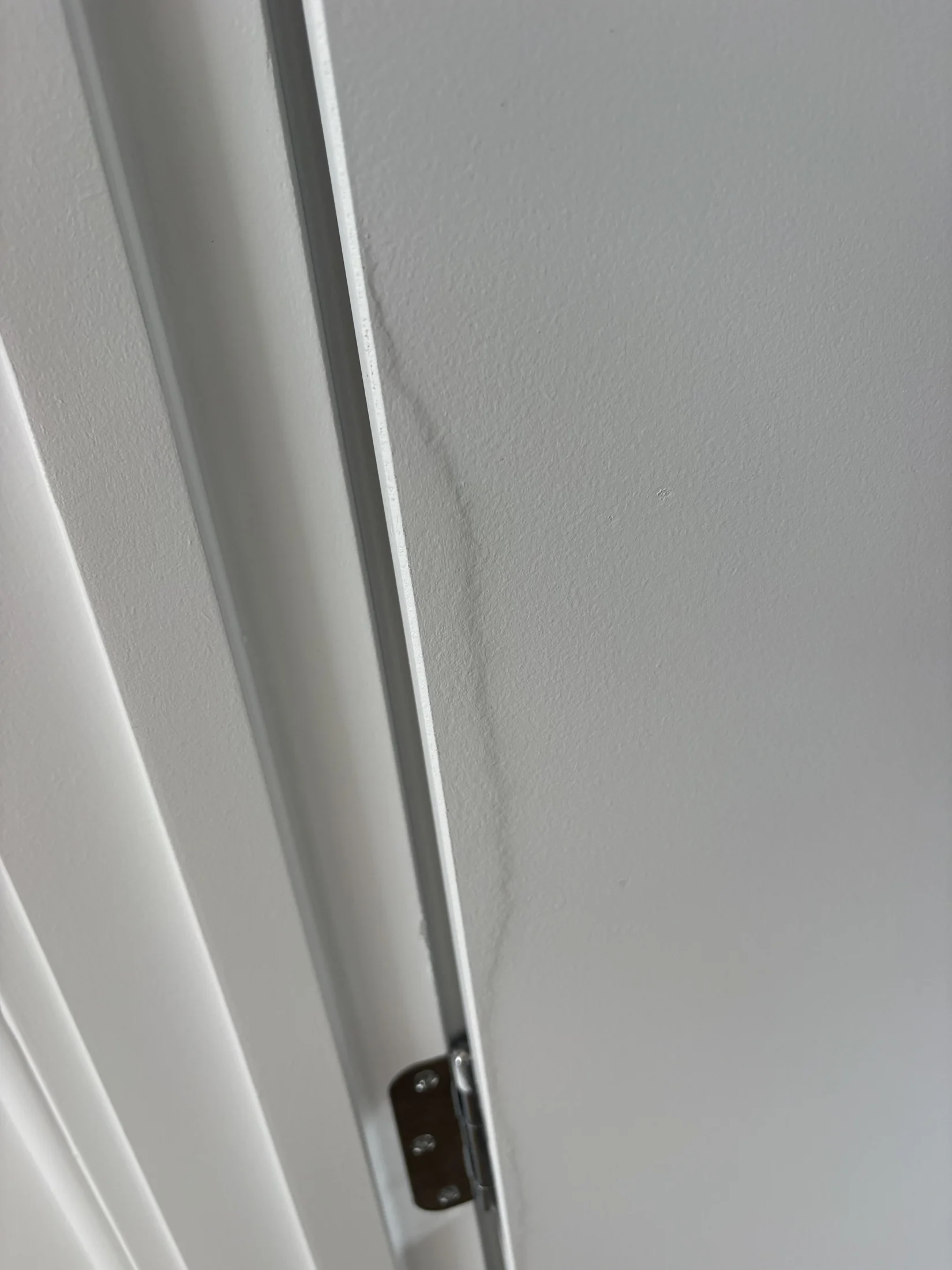 Close-up of a white door with a visible hinge and slight indentation on the surface. The background is a plain white wall, creating a minimalistic feel.