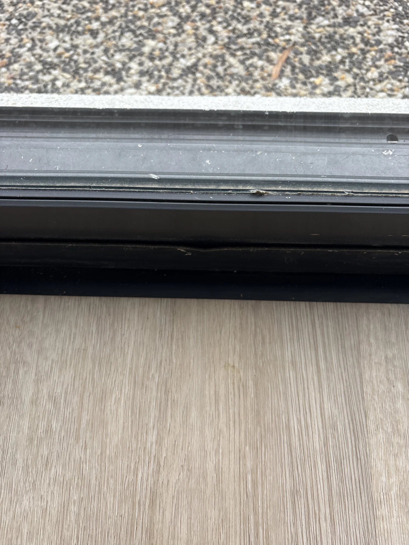 Close-up of a dusty window sill with a wooden floor beneath and a pebble-textured surface outside, creating a neutral, calm mood.