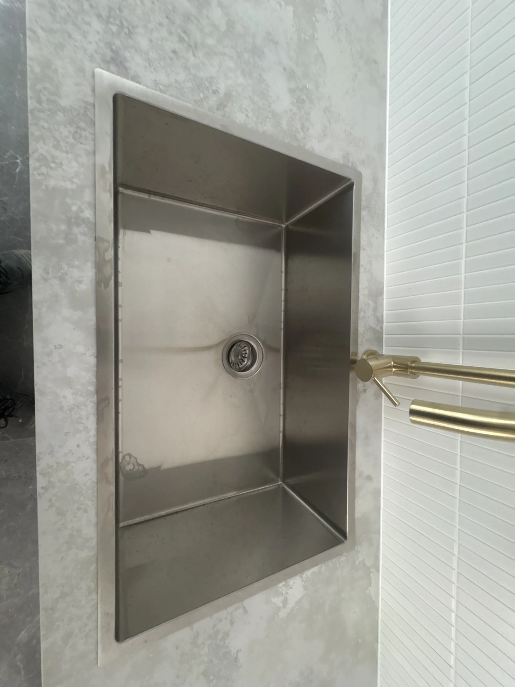 Stainless steel sink with gold faucet on a gray marbled countertop. White textured wall panel in the background. Modern kitchen design.