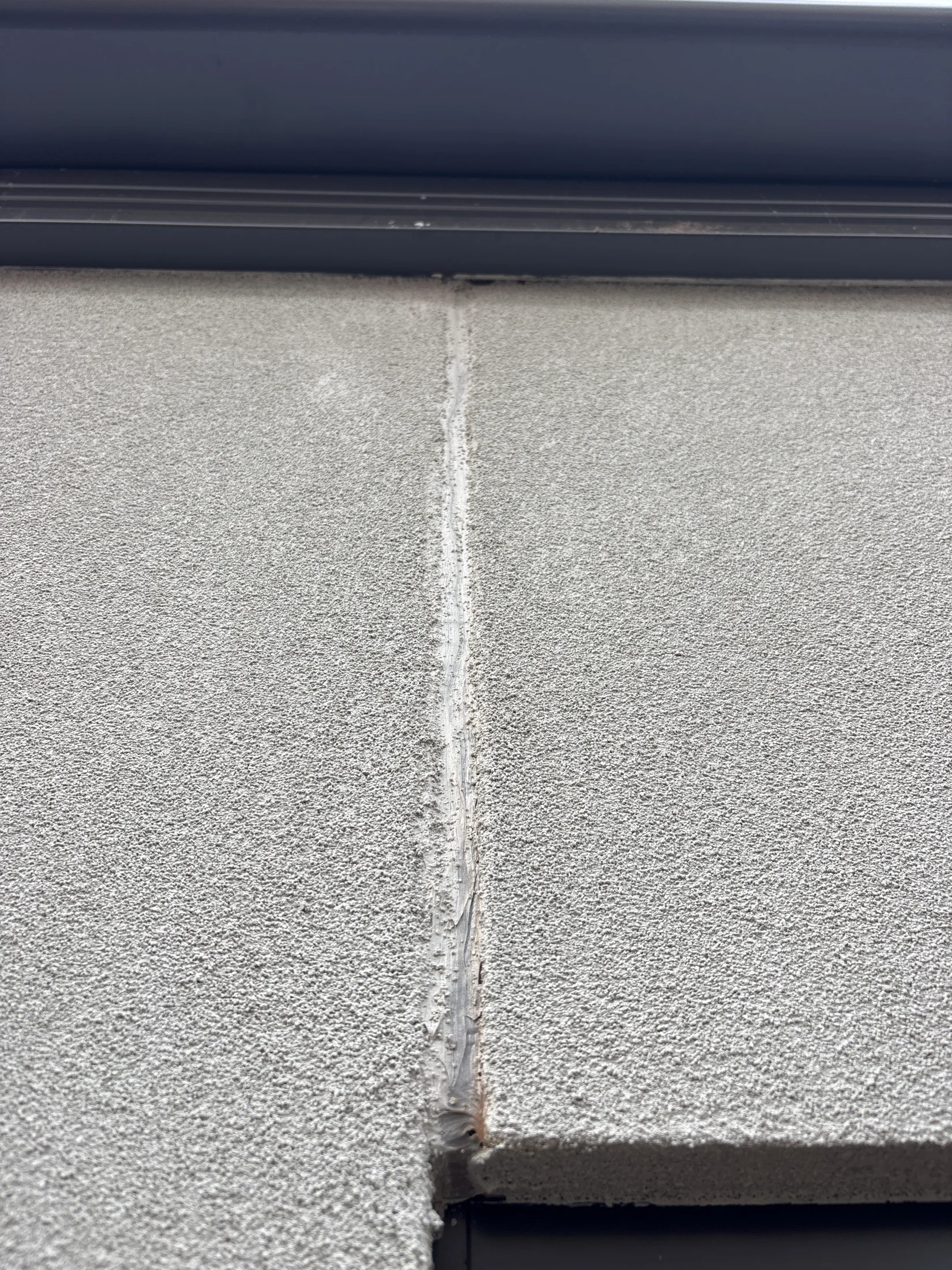Close-up of a textured concrete wall with a vertical sealed joint. Light gray color, dark trim at the top, no text or notable actions.