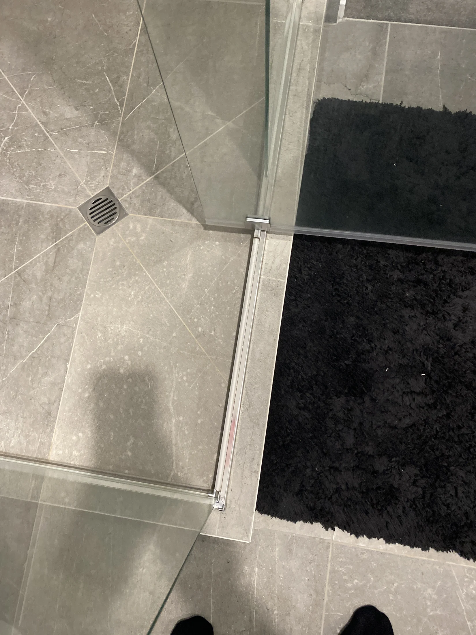 Glass shower corner with gray tiled floor and black rug. A silver drain is visible, and a faint shadow is cast on the tiles.