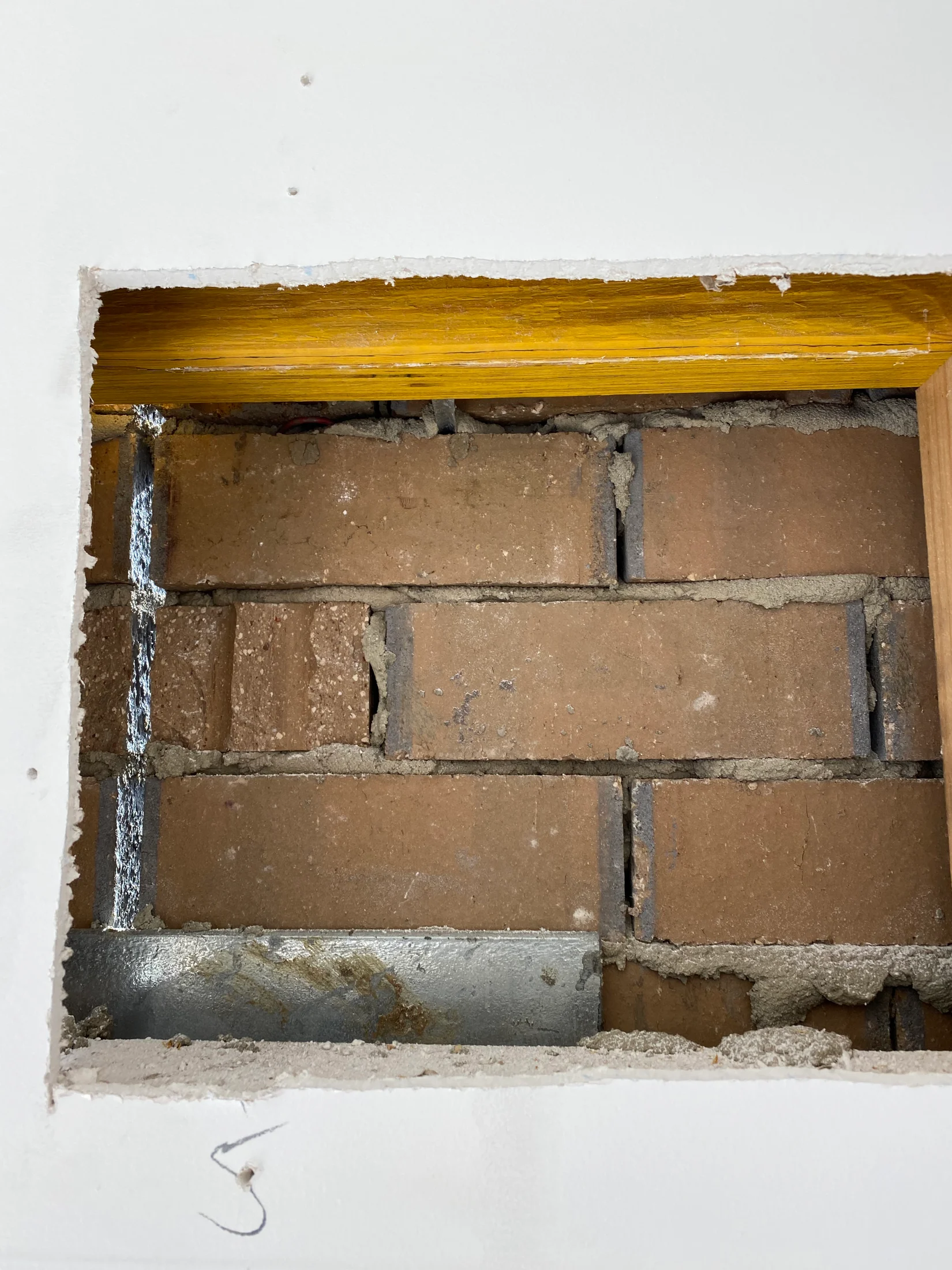 Square hole in drywall reveals red brick wall behind it. A yellow wood beam is visible at the top. Number "5" is written on the wall.