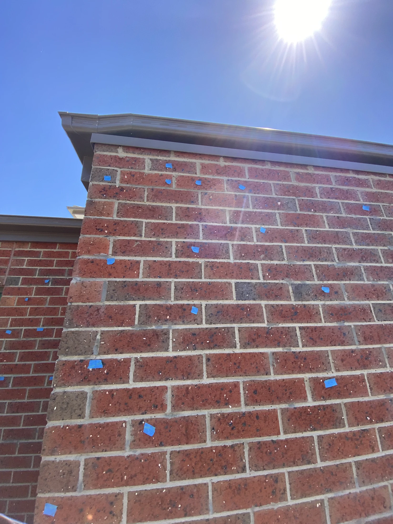 Sunny day, red brick wall with blue tape marking spots, against a clear blue sky. Sunlight creates bright reflections on the wall.
