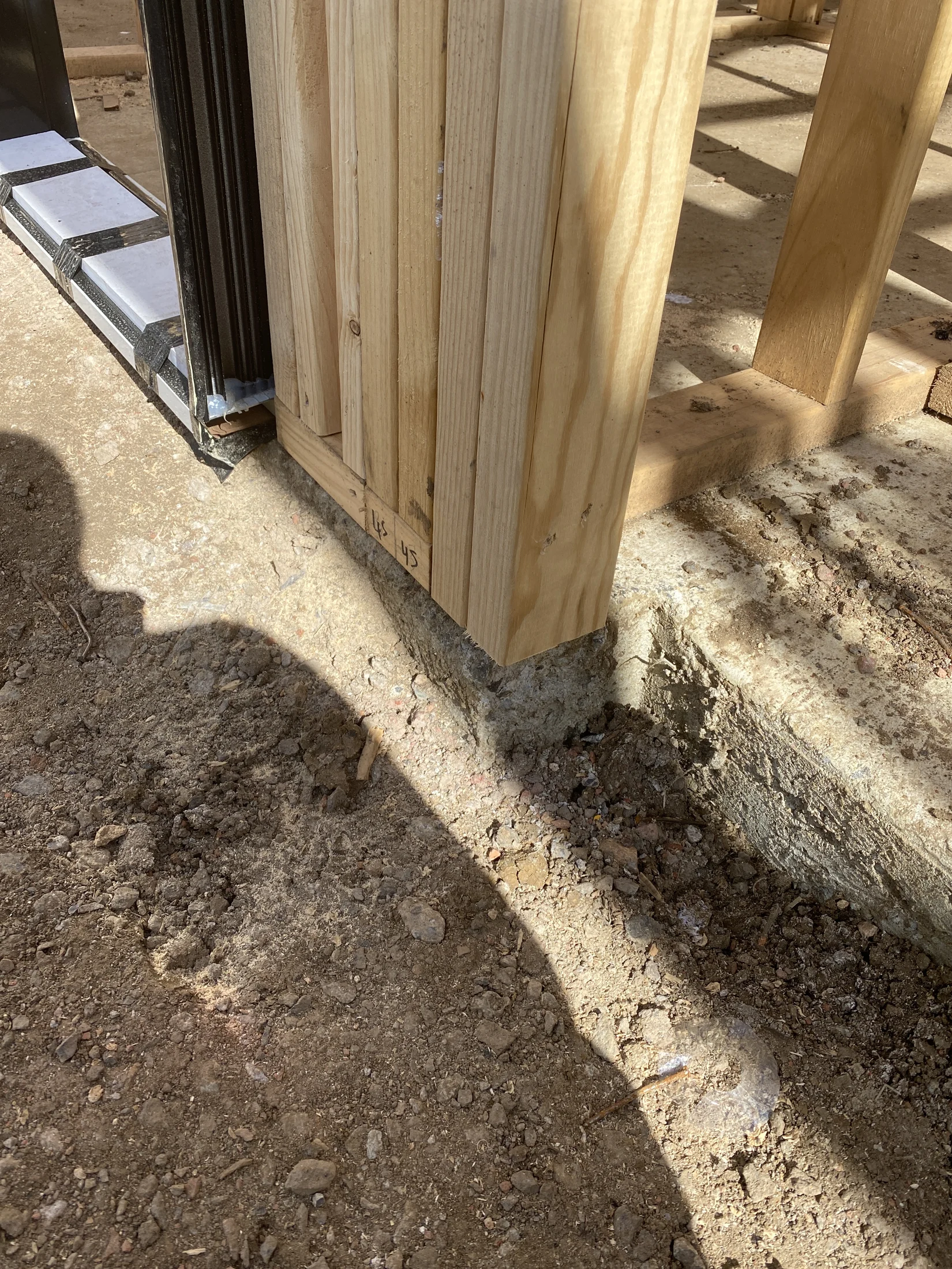 Wood frame and metal support on a dirt floor at a construction site. Shadows and sunlight create contrast. Numbers written on wood.