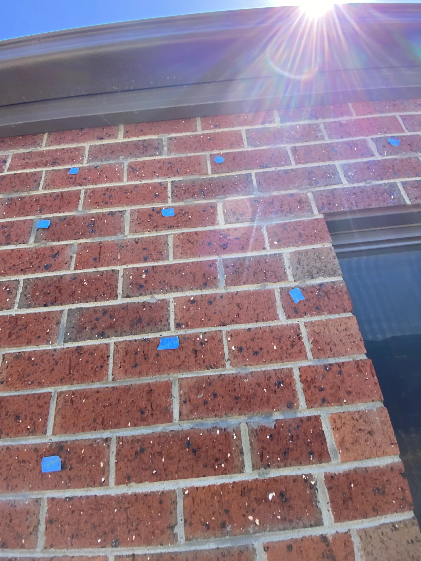 Red brick wall with scattered blue tape pieces under a bright sunlit sky. Sun rays create a lens flare effect. No visible text.