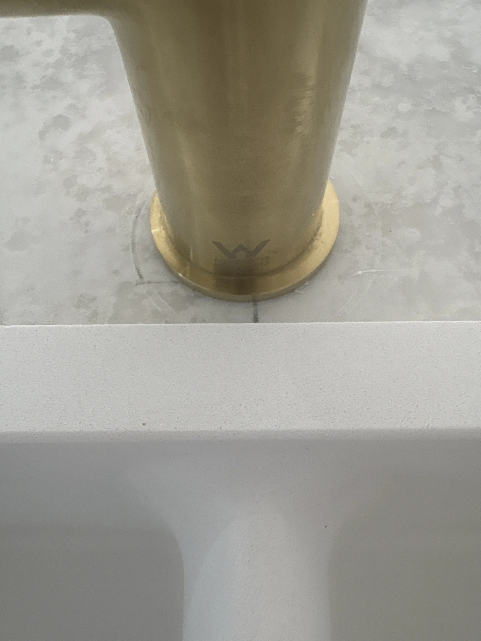 Golden metallic faucet base on a speckled gray surface with a visible "W" logo. White countertop partially in view, creating a clean aesthetic.
