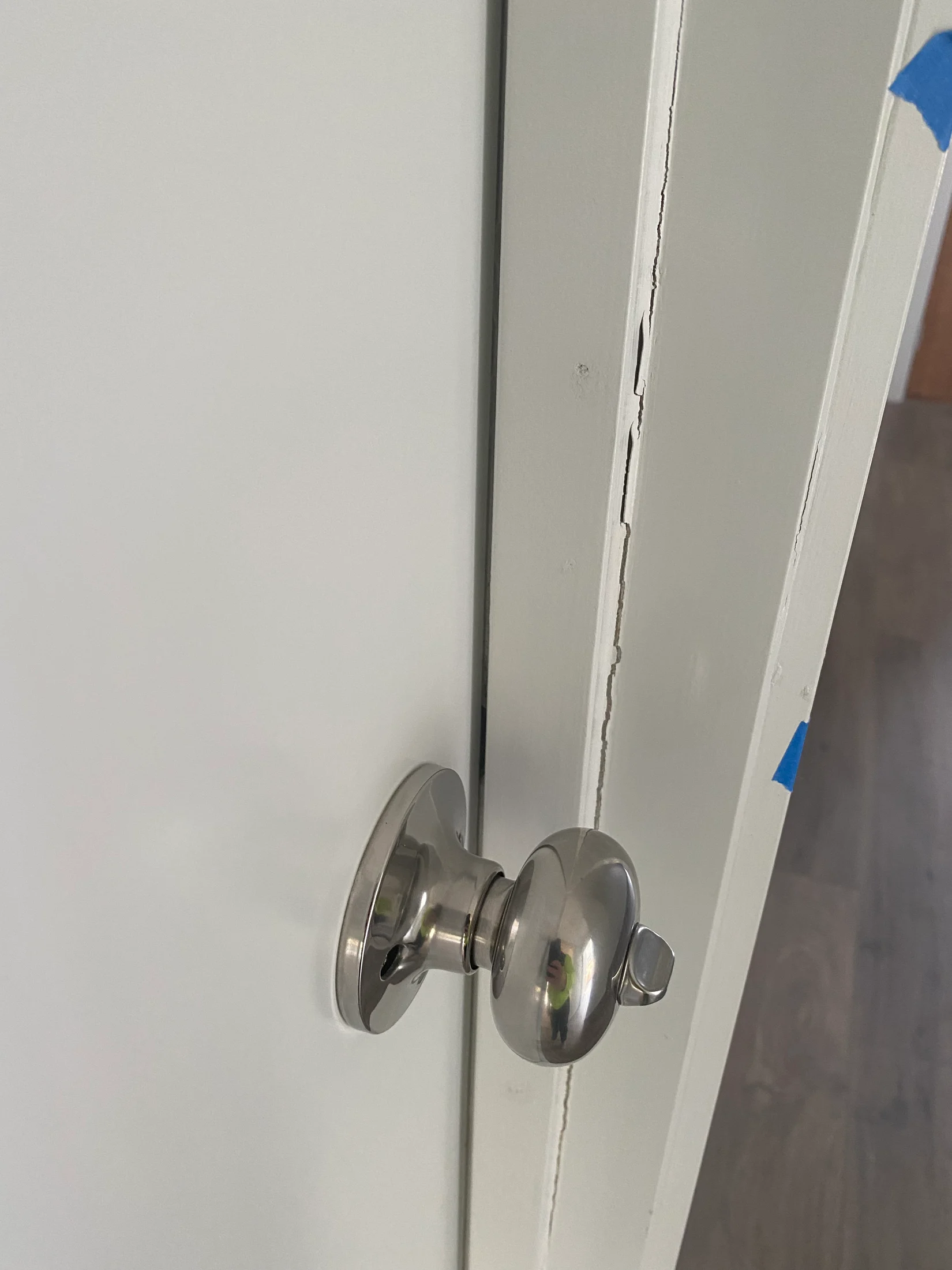 Silver doorknob on a white door slightly ajar, with a visible paint chip and blue tape on the frame, revealing a wooden floor background.