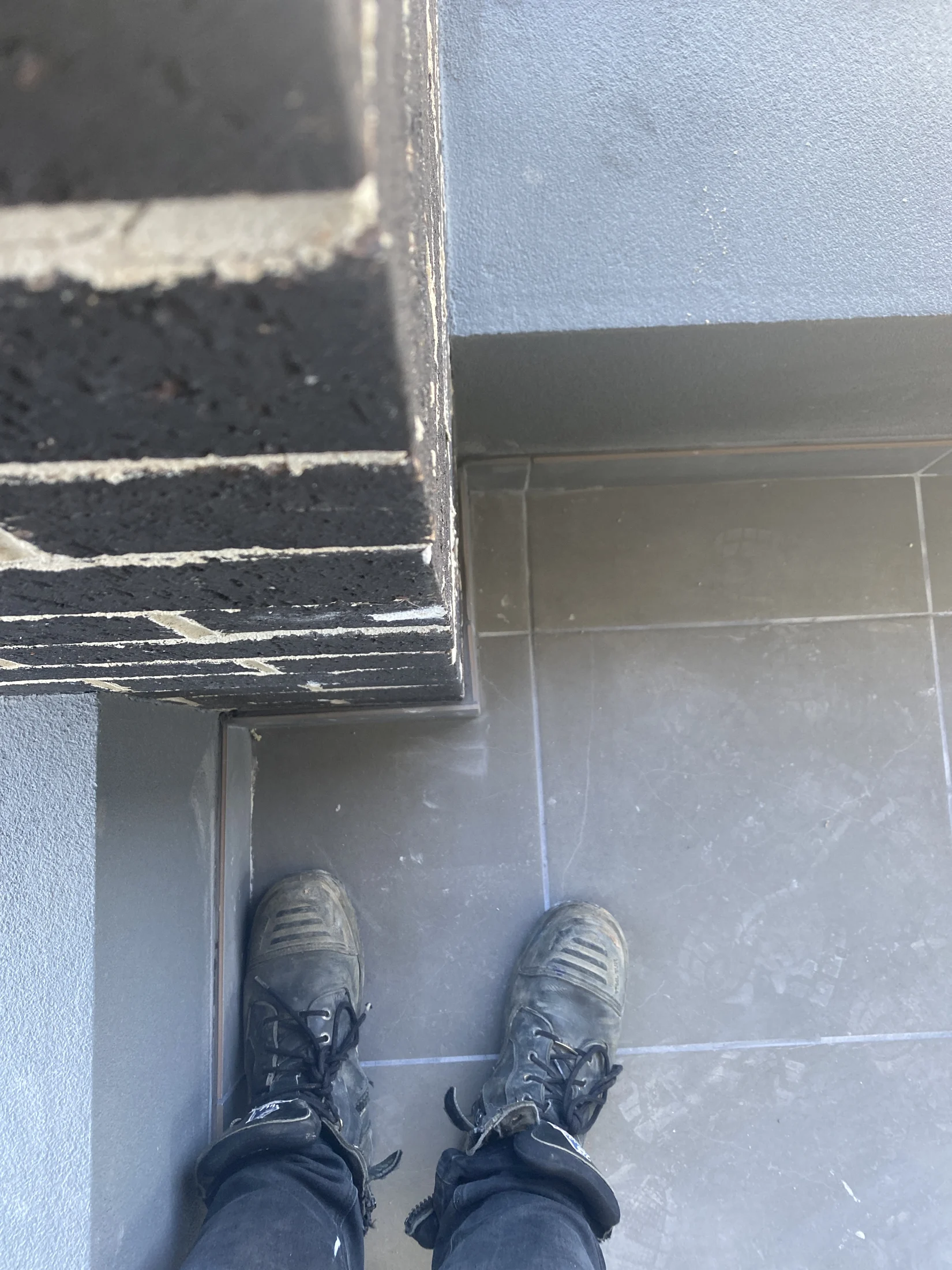 Worn black boots on gray tiled floor, next to a black brick wall. The setting appears industrial or construction-related.