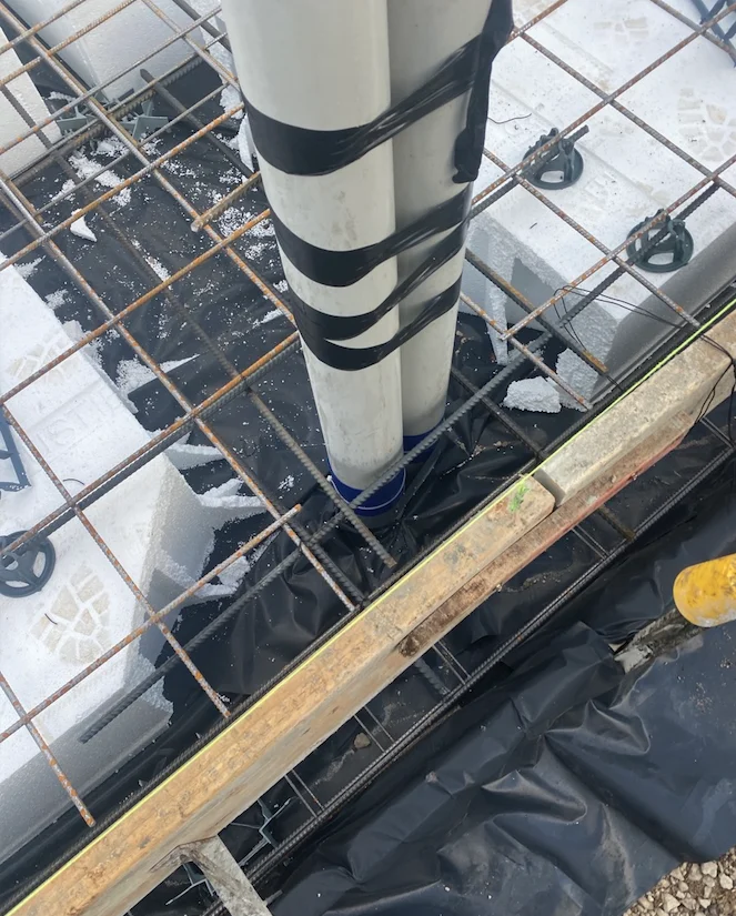 Two PVC pipes secured with black tape within a metal grid frame on a construction site. White foam blocks and black plastic are visible.