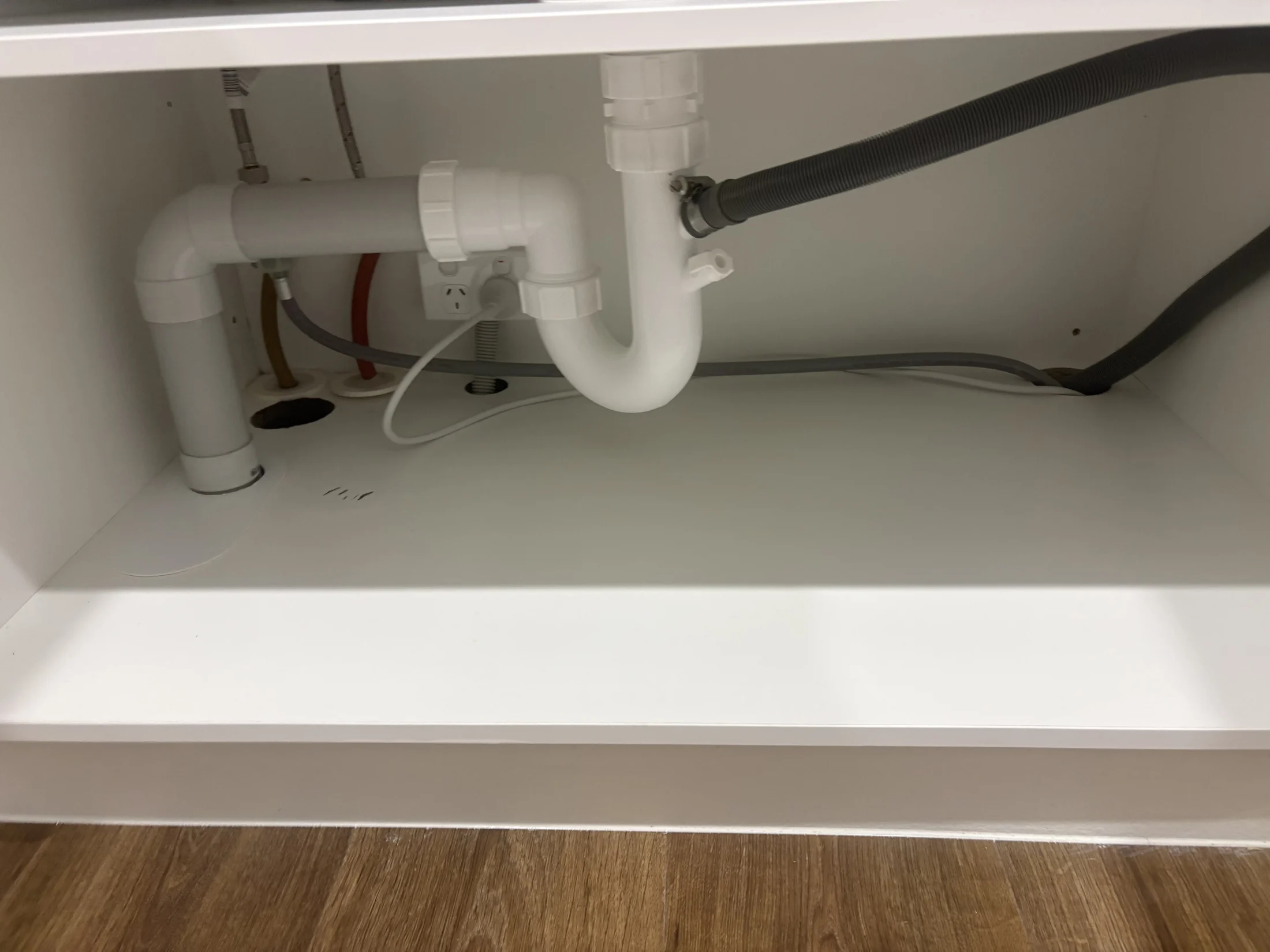 Cabinet interior with white plumbing pipes, a plug socket, and hoses. Wooden floor visible below, with a clean, organized appearance.