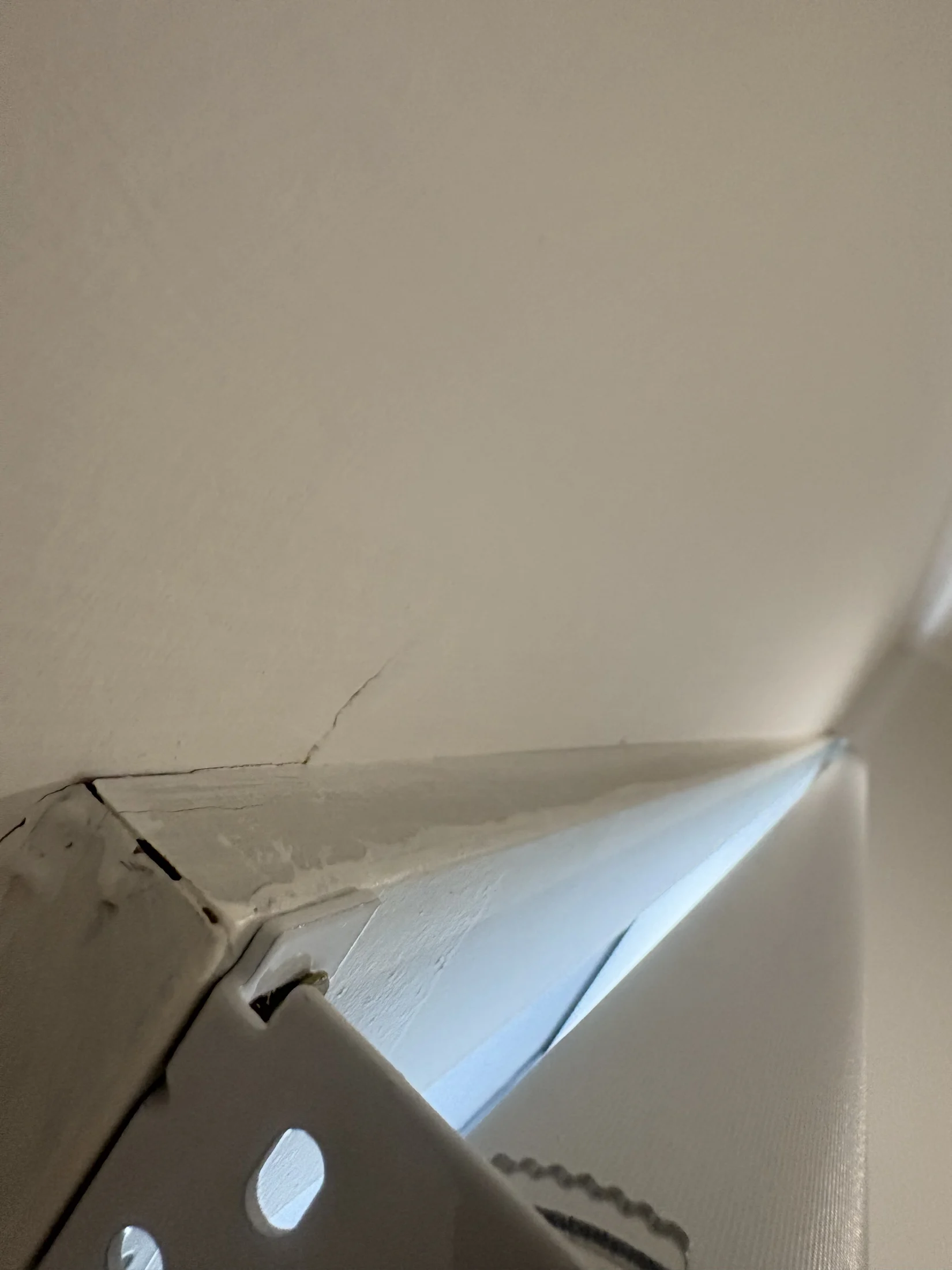 Close-up of a cracked white ceiling corner with a visible gap, showcasing light blue sky through the opening. Slight mood of disrepair.