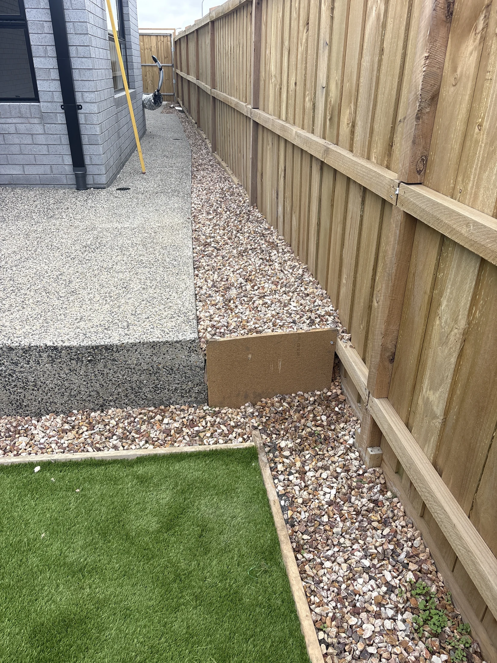 A narrow backyard with a wooden fence features a gravel path, concrete steps, and a patch of artificial green grass. A ladder leans on a gray brick wall.