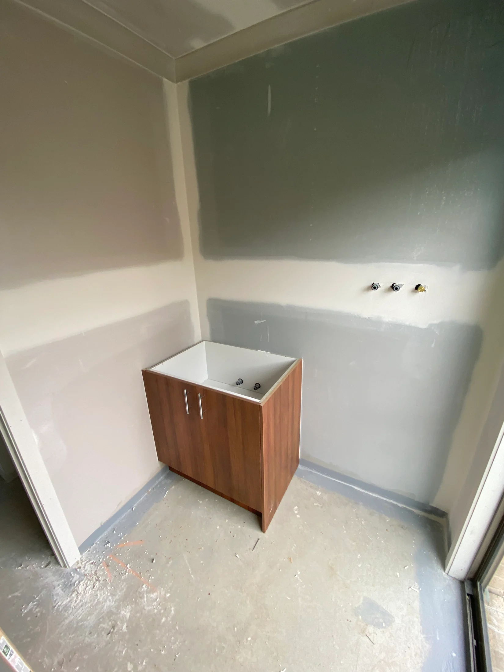 Unfinished room with gray walls, a wooden cabinet with a sink basin, and exposed plumbing. Concrete floor, minimalistic setting.
