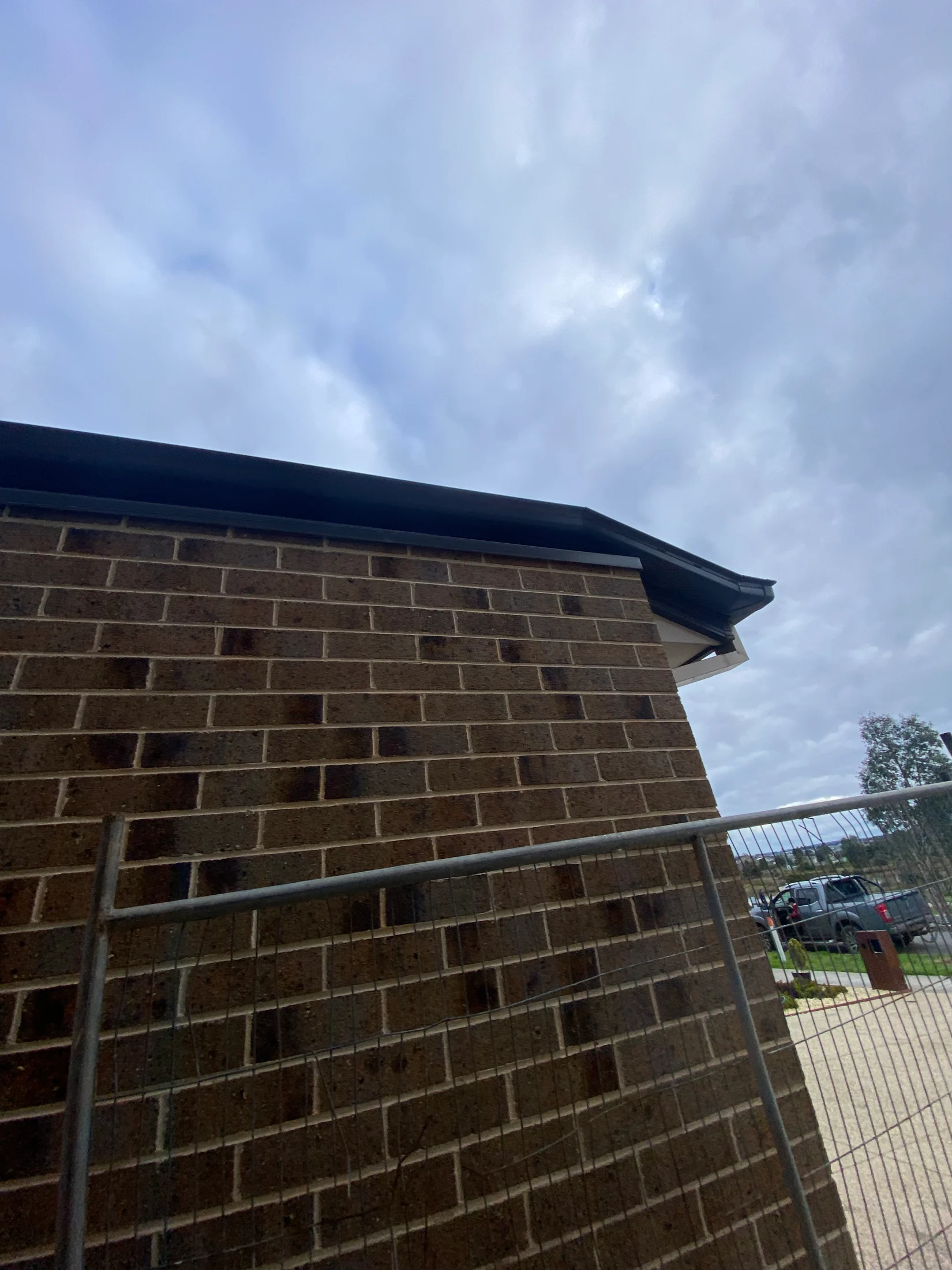 Brick wall and metal fence under a cloudy sky. A car and trees are visible in the distance, creating a calm, overcast mood.