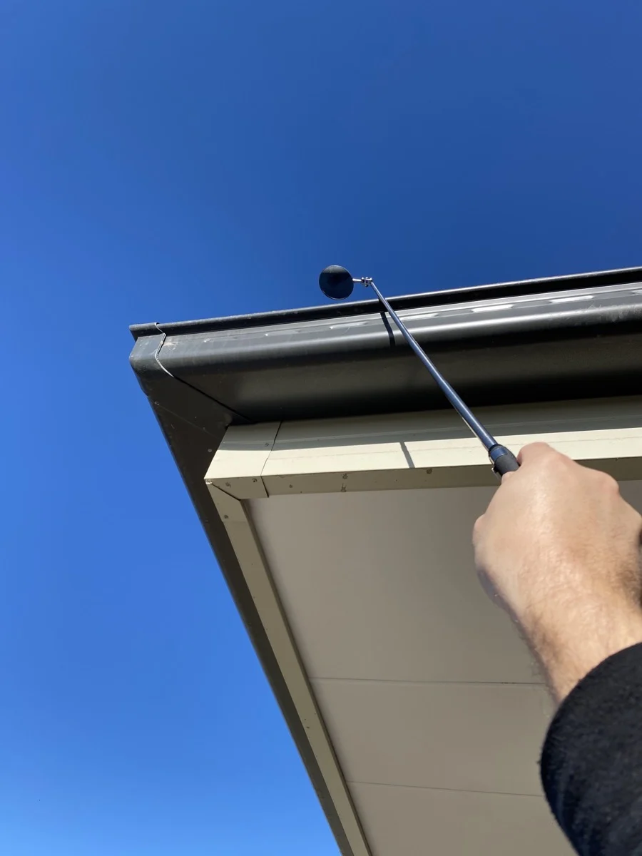 Hand holds a black sphere on a pole, reaching towards a rooftop under a clear blue sky. The building's edge is visible at an angle.