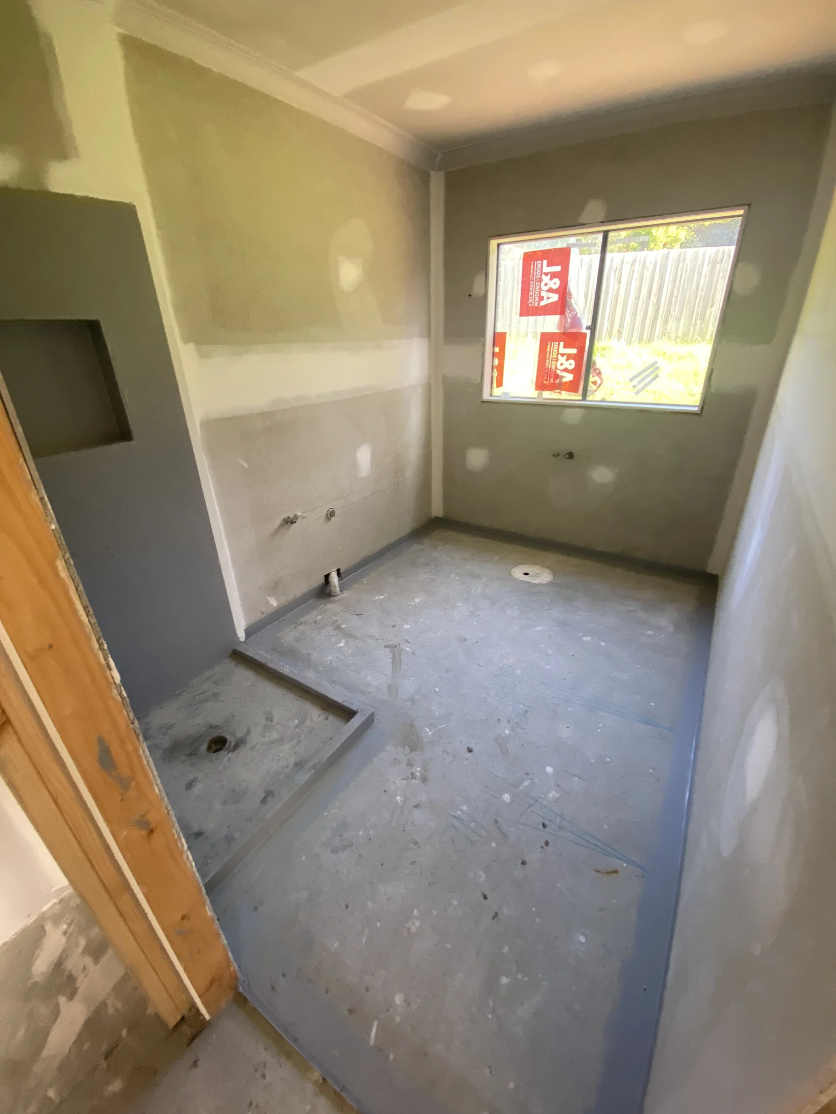 Unfinished bathroom under construction with drywall and cement floor. Window with red text, sunlight streaming in. Neutral colors.