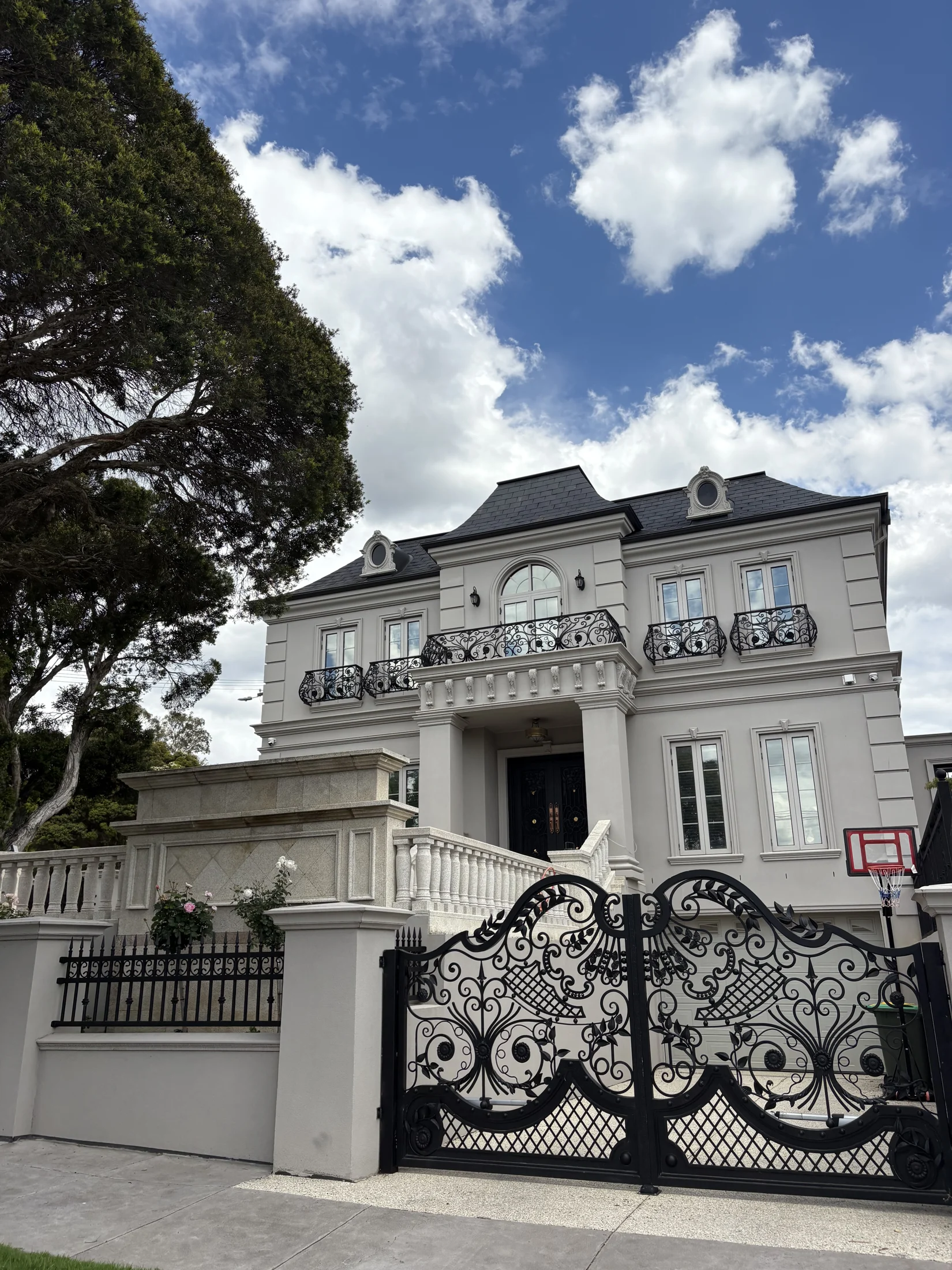 Elegant gray mansion with ornate black gate, white stone staircase, and basketball hoop. Blue sky with clouds in the background.