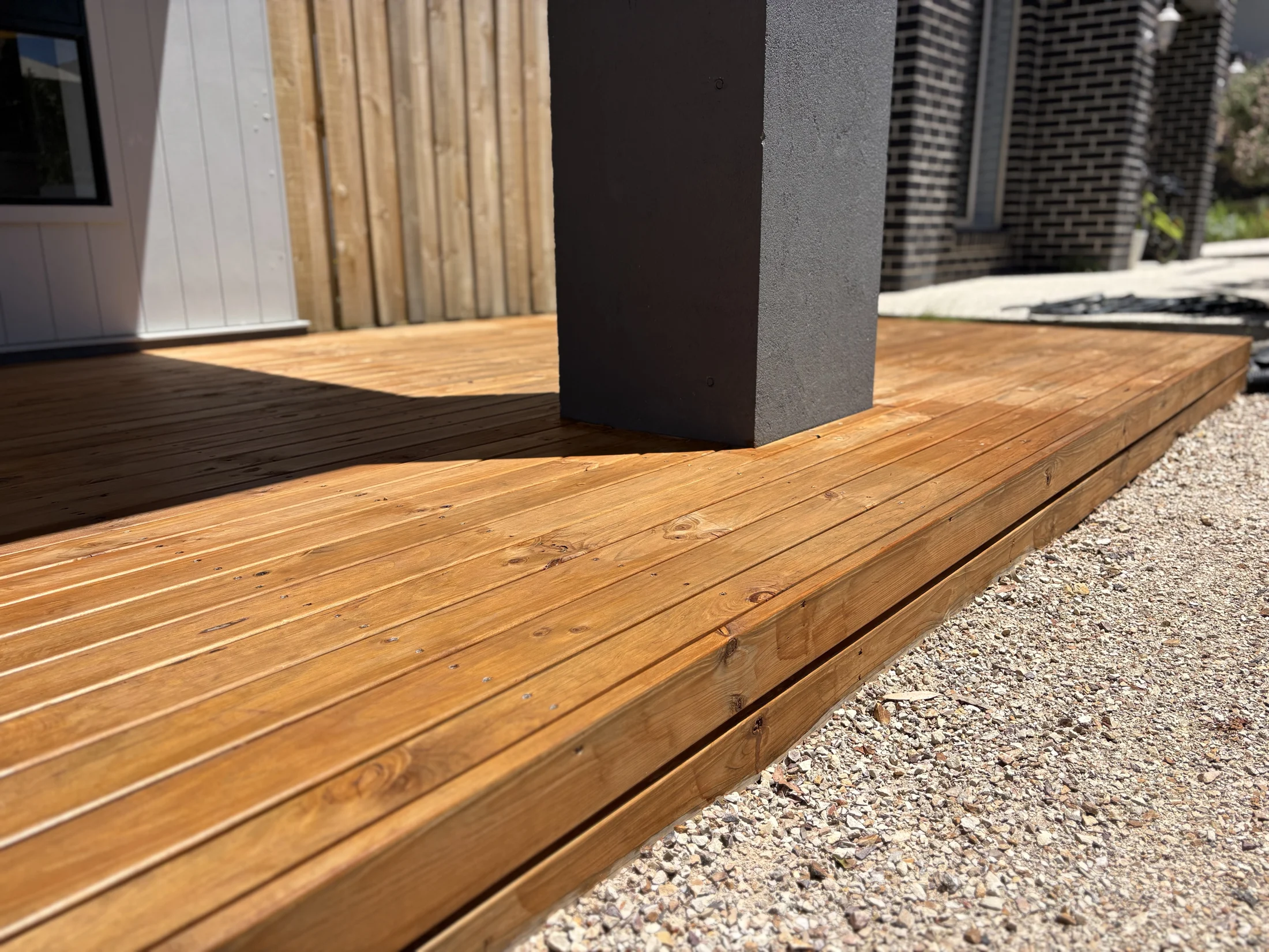 Wooden deck with a gray pillar, surrounded by brick and wood walls. Sunlight casts shadows on the deck. Pebbled ground nearby.