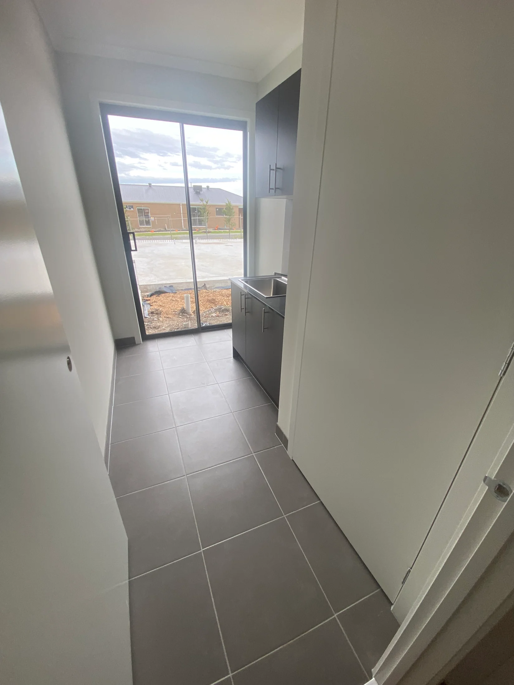 Narrow laundry room with gray tiles, dark cabinets, and a stainless steel sink. Glass door opens to a construction site under a cloudy sky.