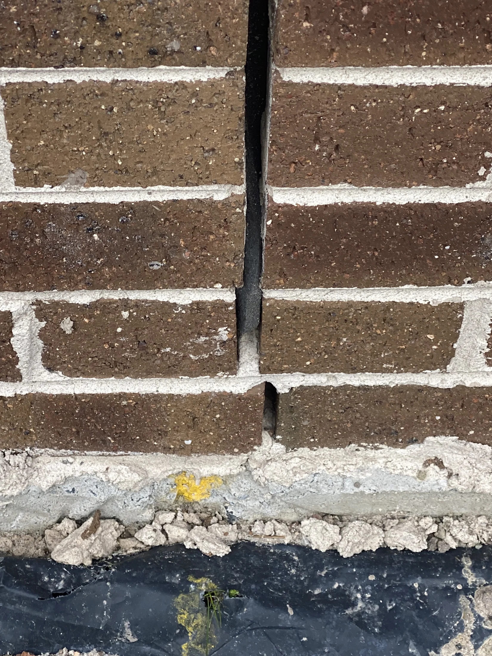 Close-up of a brick wall with a vertical crack and exposed mortar. Dark brown bricks, yellow paint mark on the foundation, black tarp below.