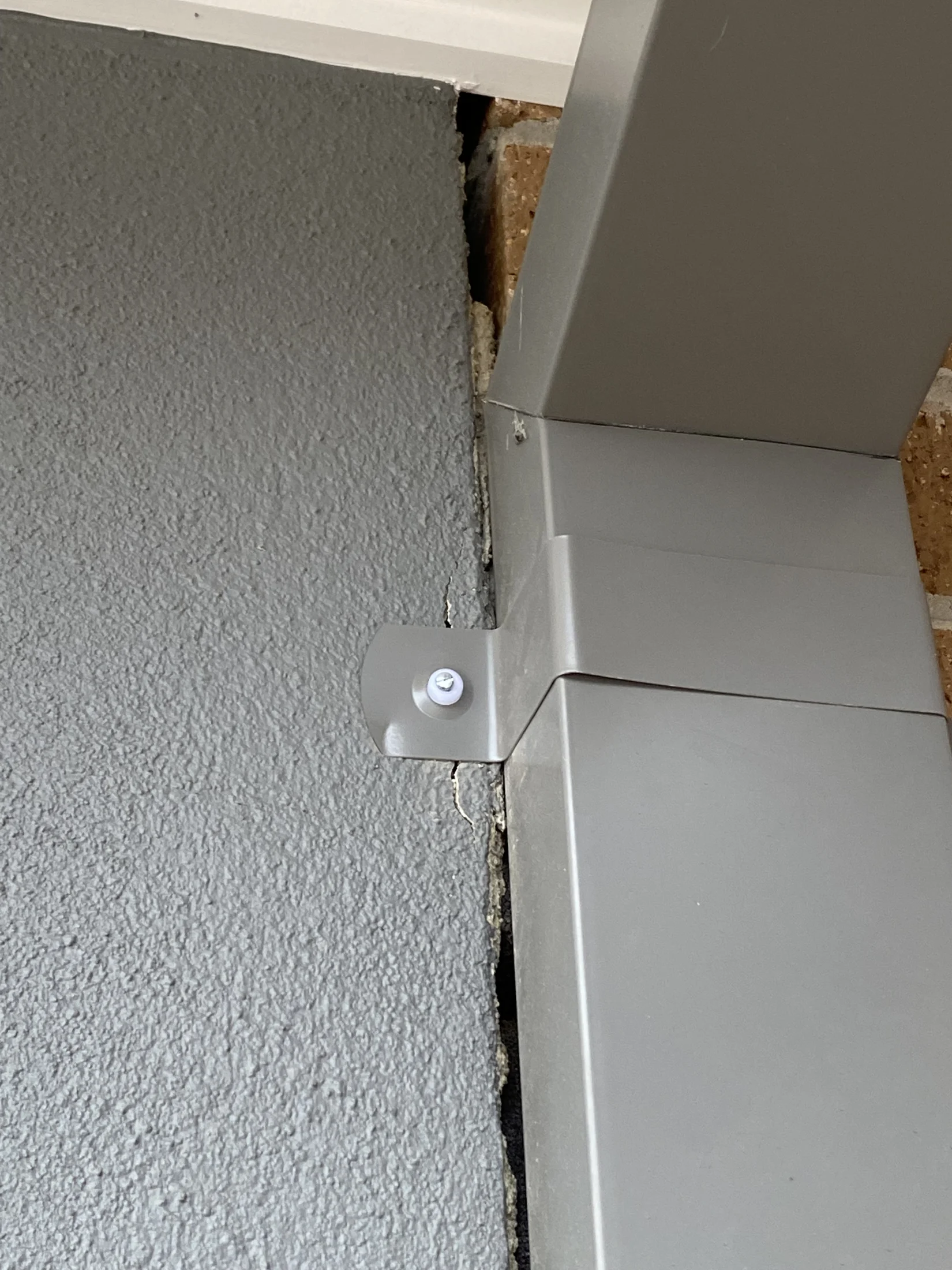 Gray wall with a metal bracket attached, showing a gap revealing bricks. The background is gray with some texture. No text visible.