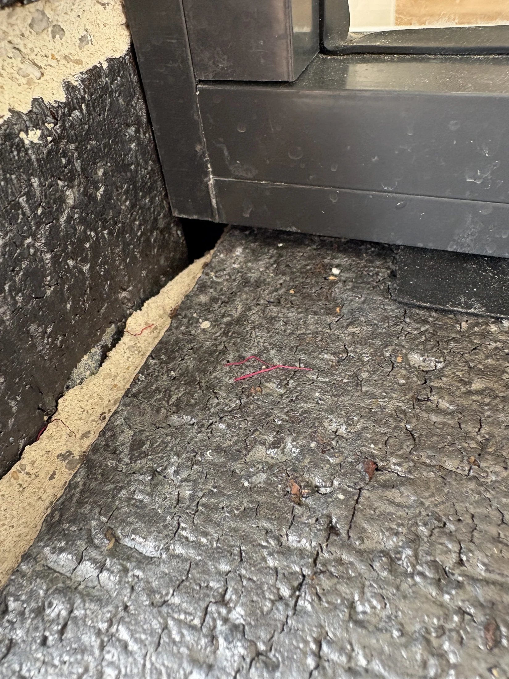 Cracked asphalt surface with a black metal frame and window in corner. Texture shows weathering, with bits of red thread scattered.