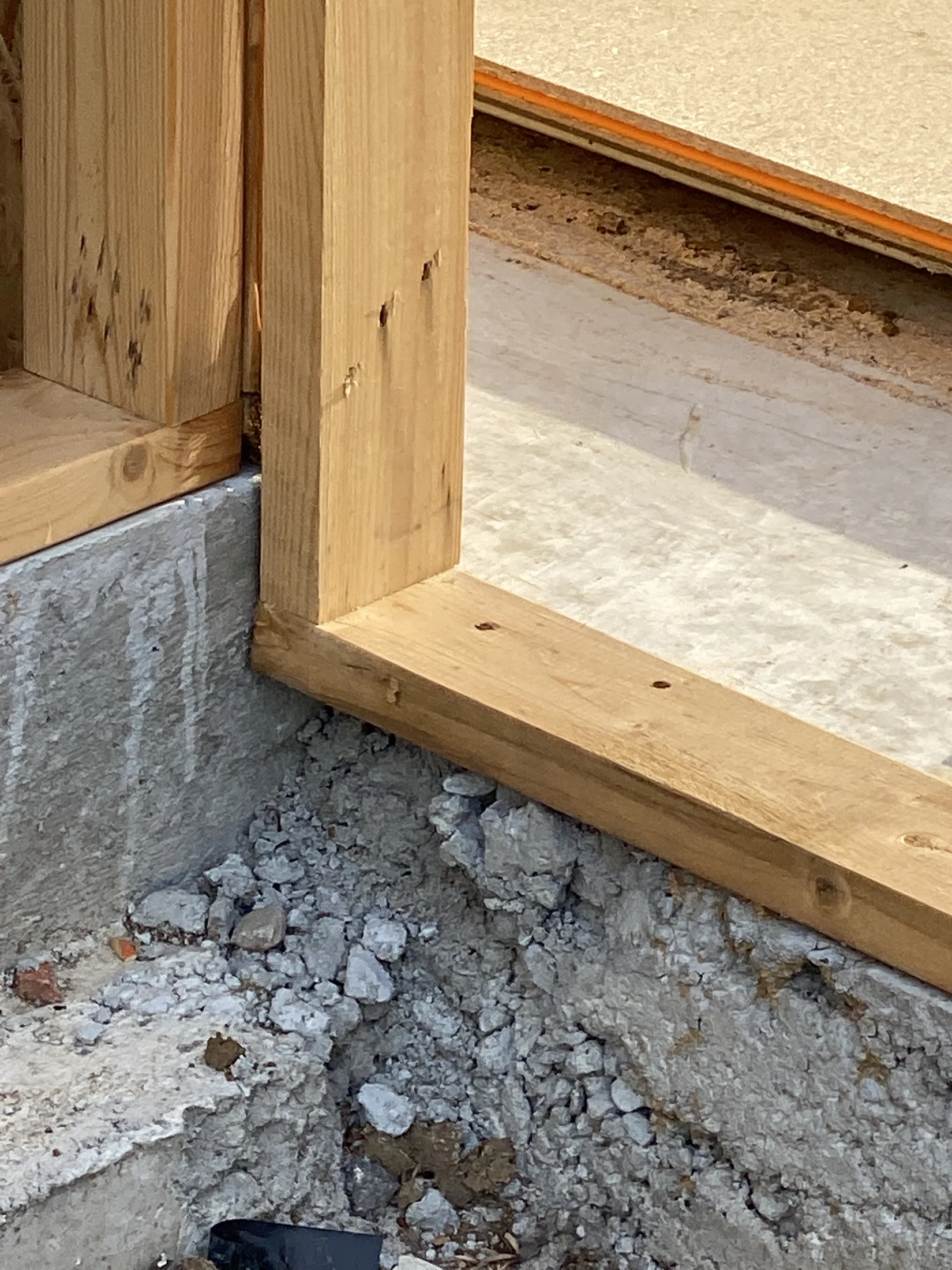 Wooden frame set on concrete, surrounded by gravel and dirt. The light-colored wood and textured concrete contrast with the rough ground.