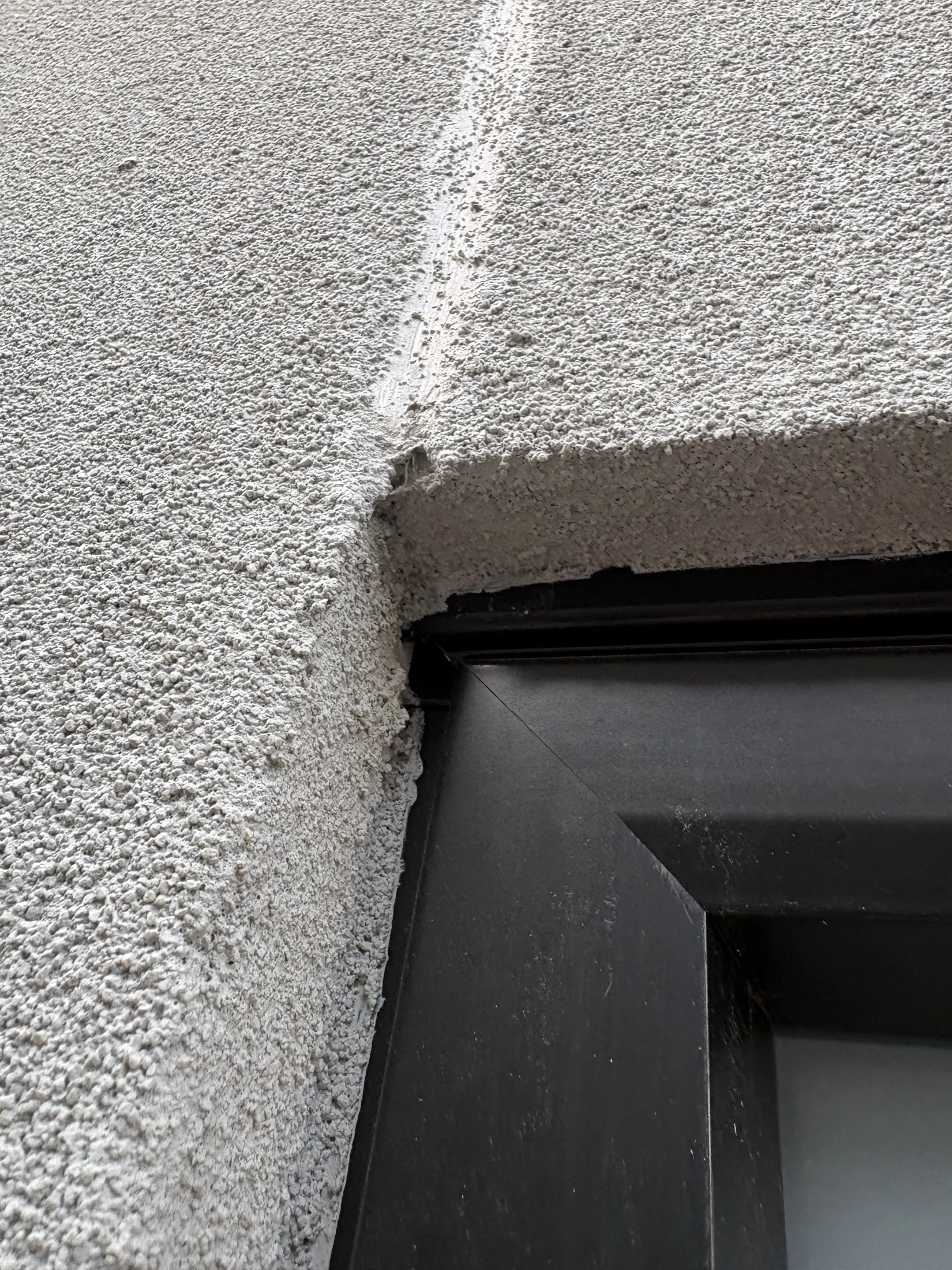 Close-up of a textured white stucco wall meeting a black window frame at a corner. The surface shows a visible seam and slight wear.