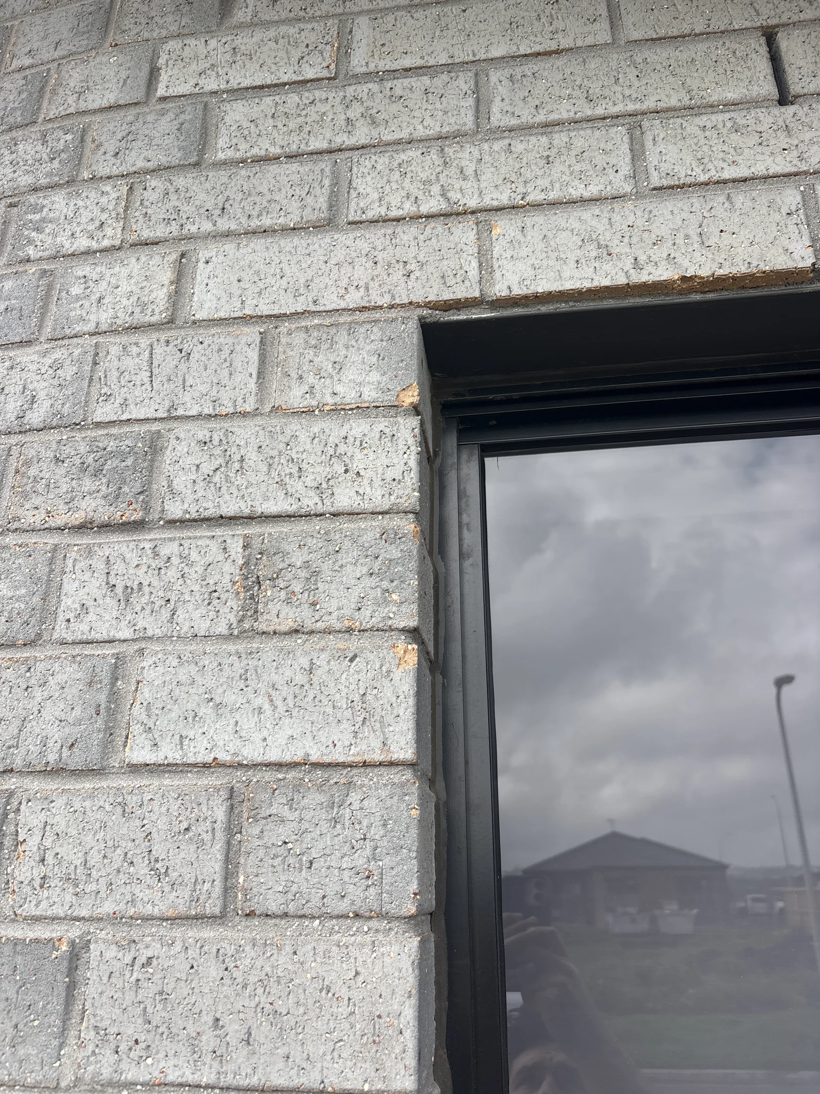 Close-up of a gray brick wall corner with visible chips near a black window. Reflective glass shows a cloudy sky and a house in the distance.