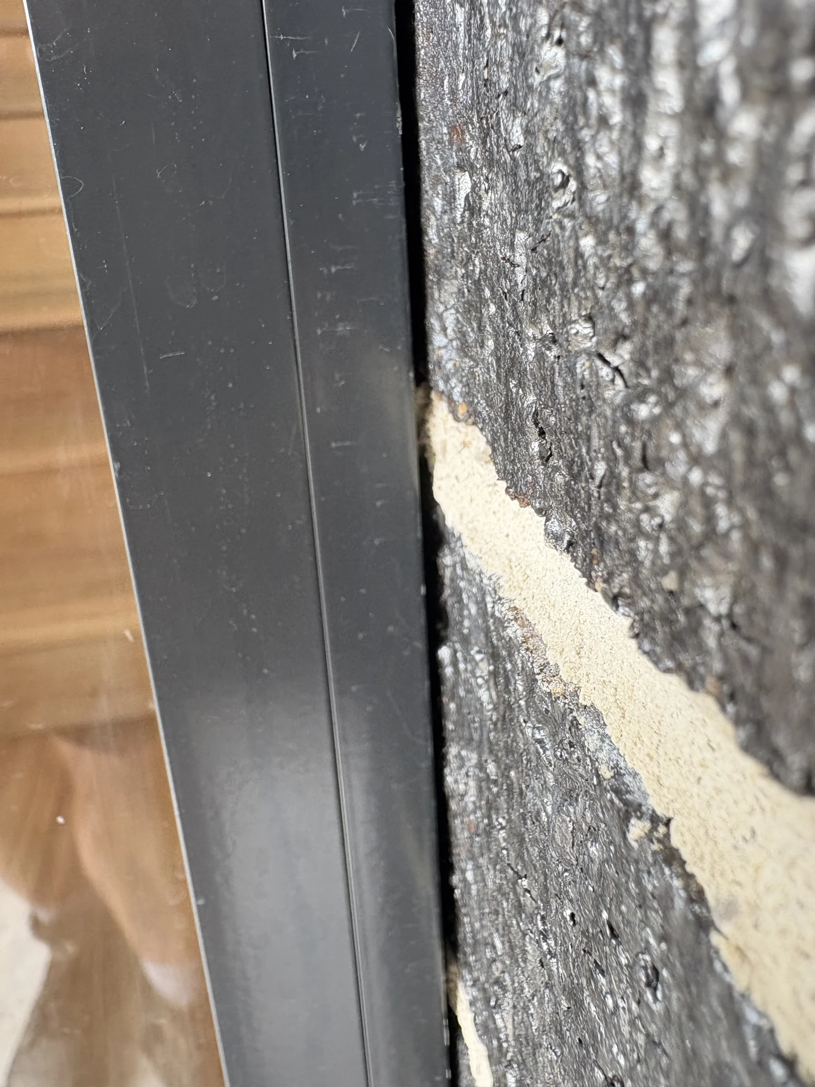 Close-up of a metal window frame next to rough, textured dark brick wall. The setting appears to be outdoors with a blurred interior view.