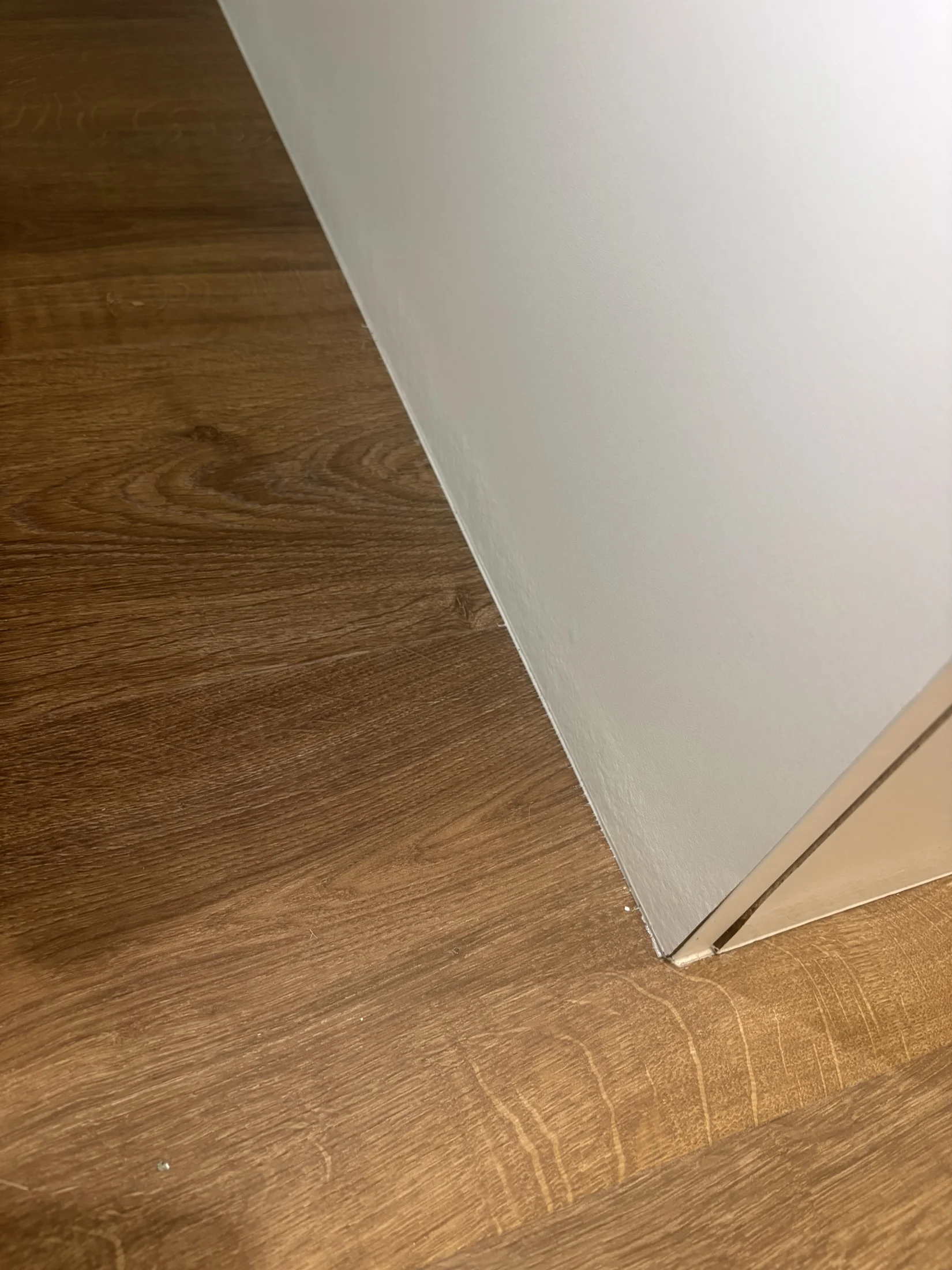 A close-up of a corner, showing a white wall meeting a wooden floor with a reddish-brown texture. The setting appears clean and simple.