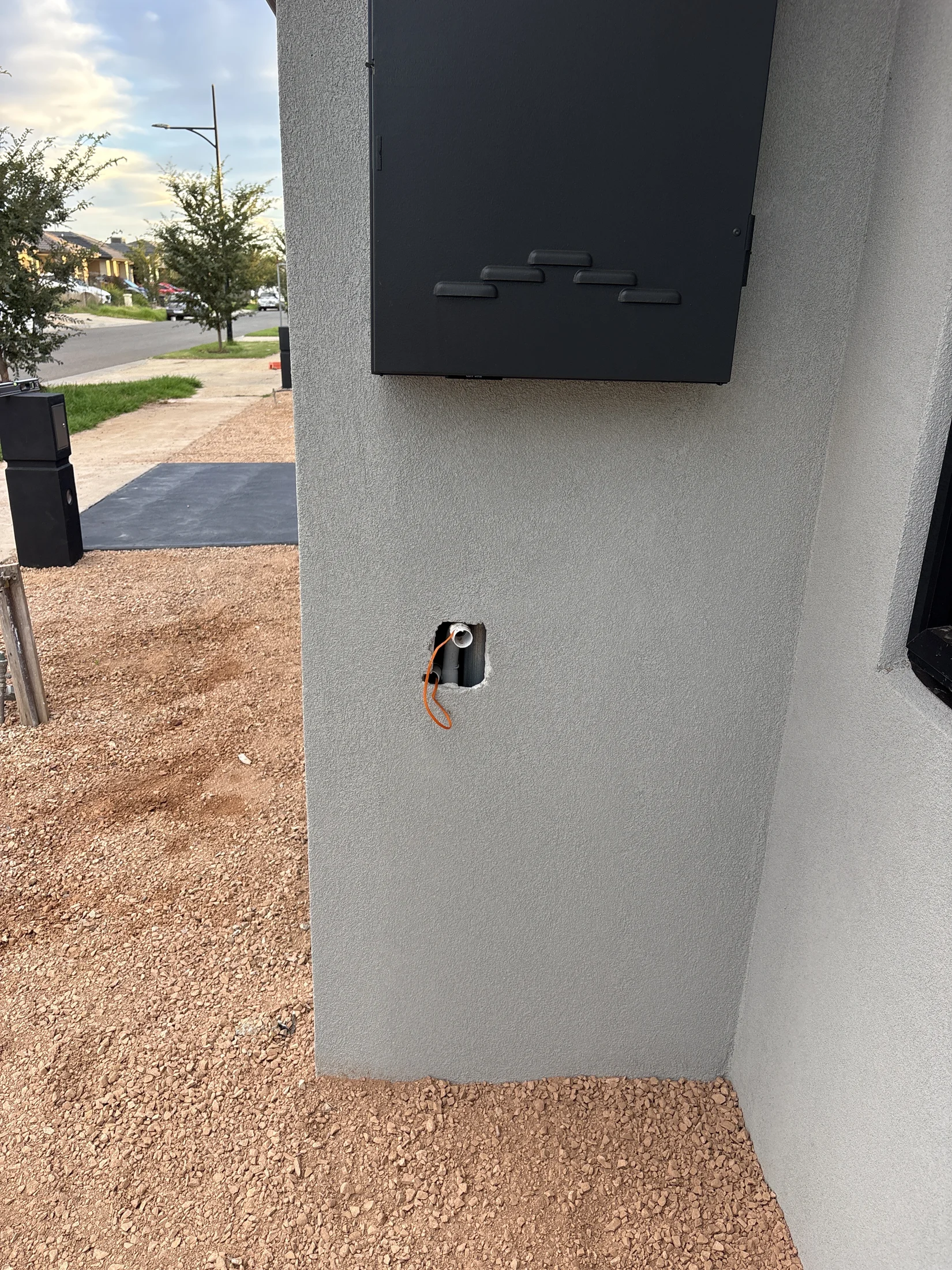 Wall with an electrical box and a hole exposing wires. Gravel ground and tree-lined street in background. Sky is partly cloudy.