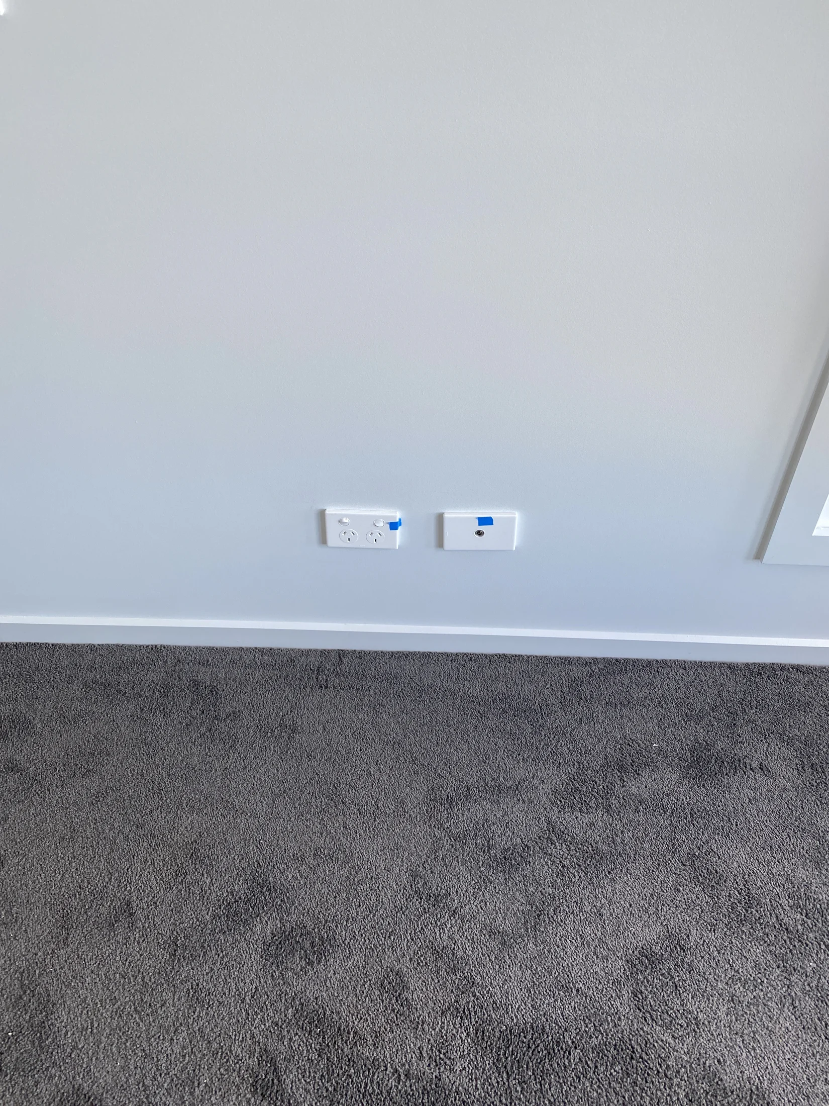 Power outlets on a light gray wall with blue tape above, over dark gray carpet. Simple and minimalistic setting, no visible text.